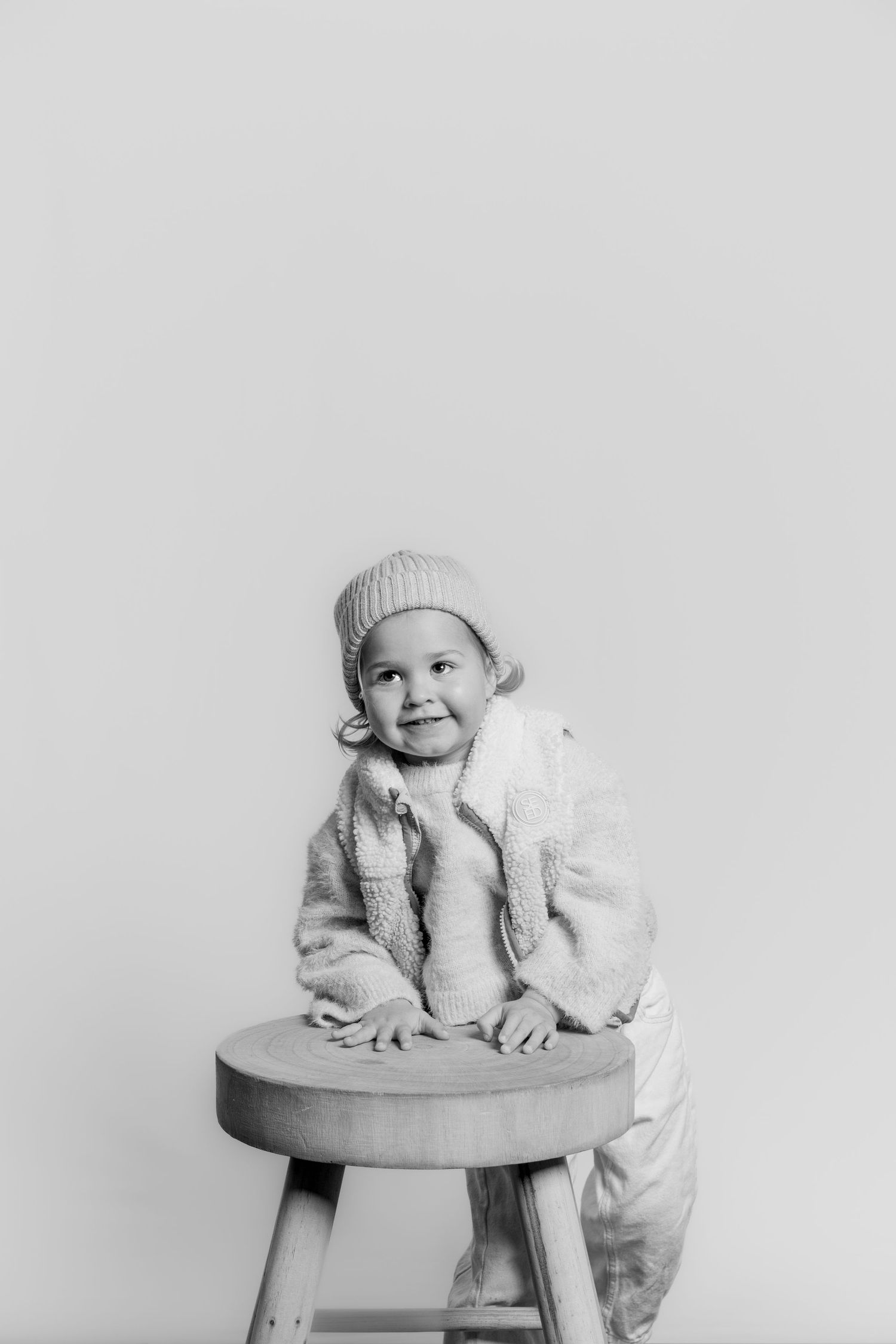 Black and white studio portrait of a happy young child in a knit hat leaning on a wooden stool and smiling brightly.