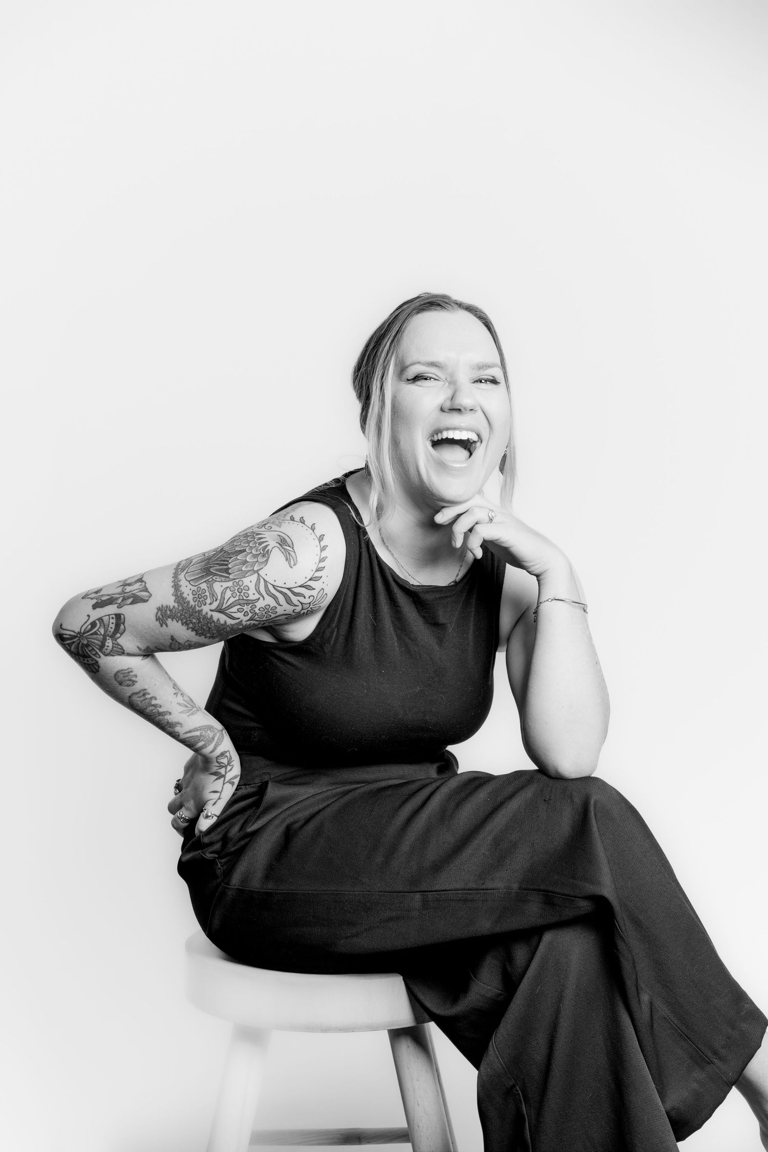 Black and white portrait of laughing tattooed woman in black outfit on studio stool.