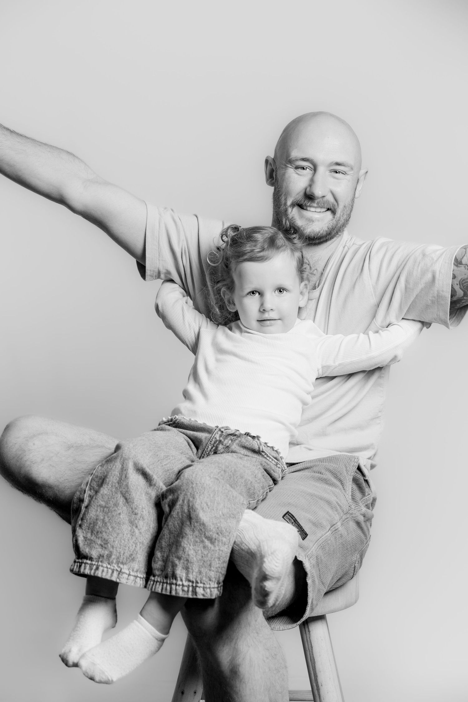 Black and white photo of a smiling bald father with tattoos holding his young daughter on his lap.