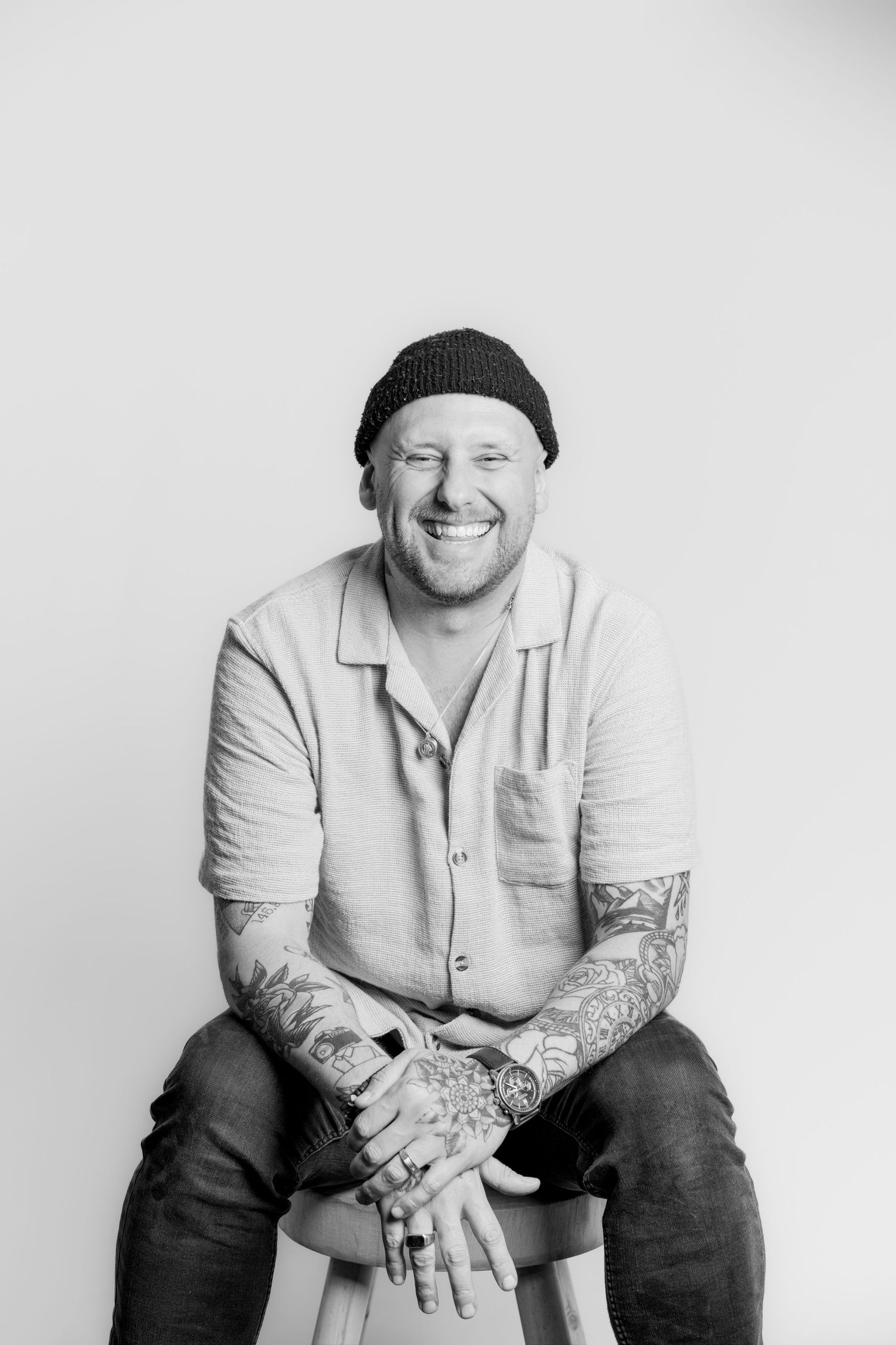 Black and white studio portrait of broadly smiling tattooed man seated on stool.