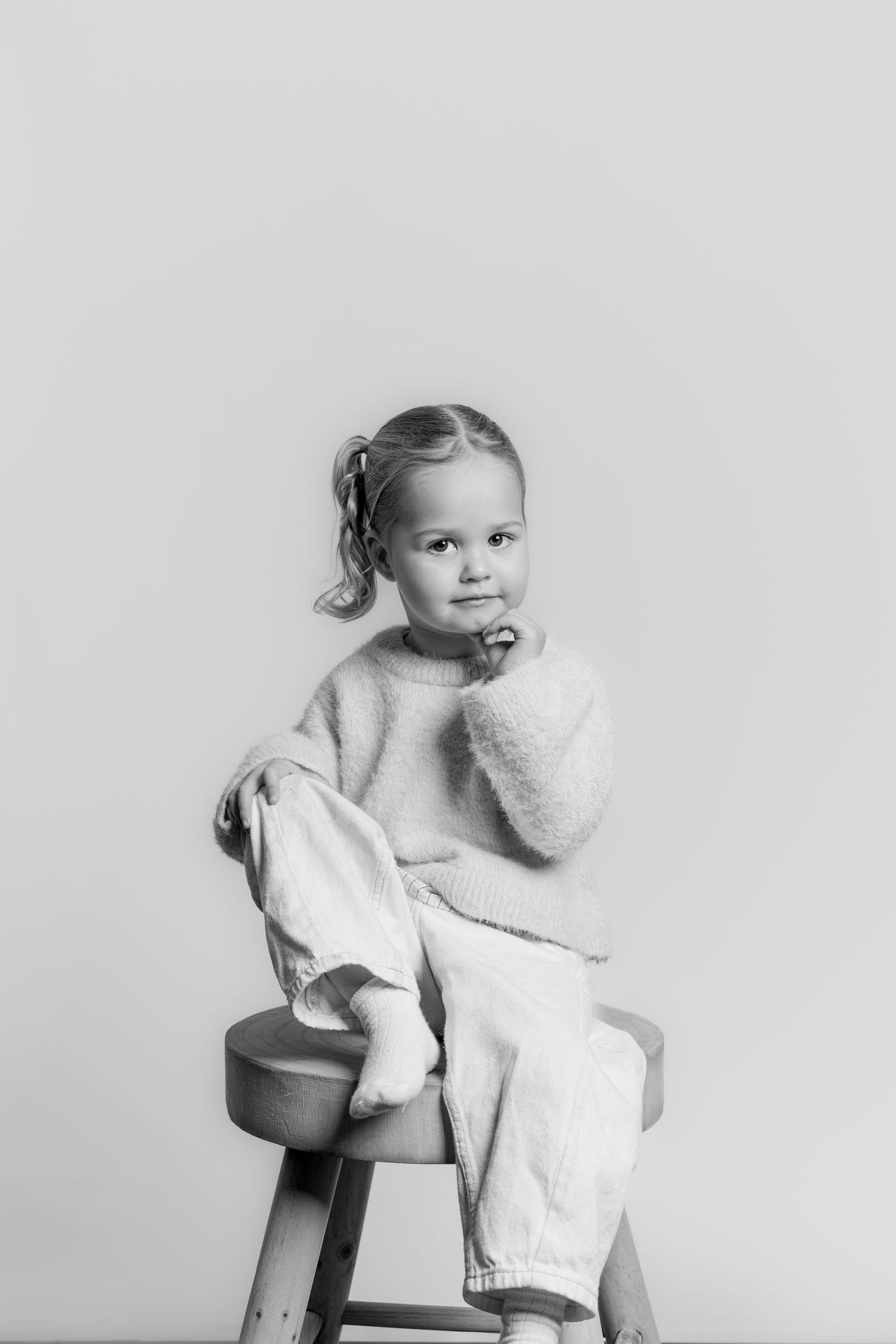 Black and white portrait of a young girl sitting on a wooden stool, resting her chin on her hand.