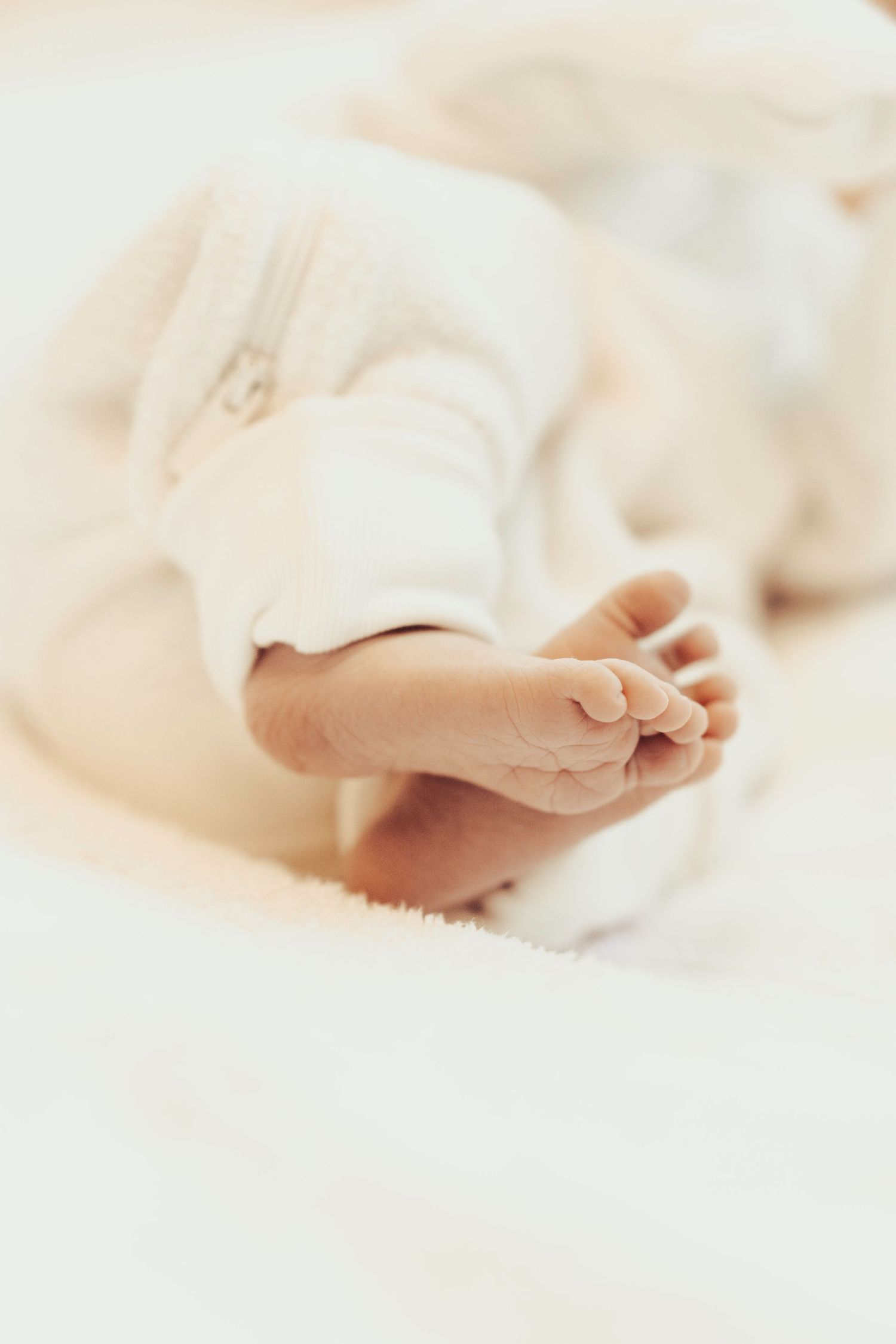 Close-up of tiny newborn baby feet in white zip-up pajamas on a soft white blanket.