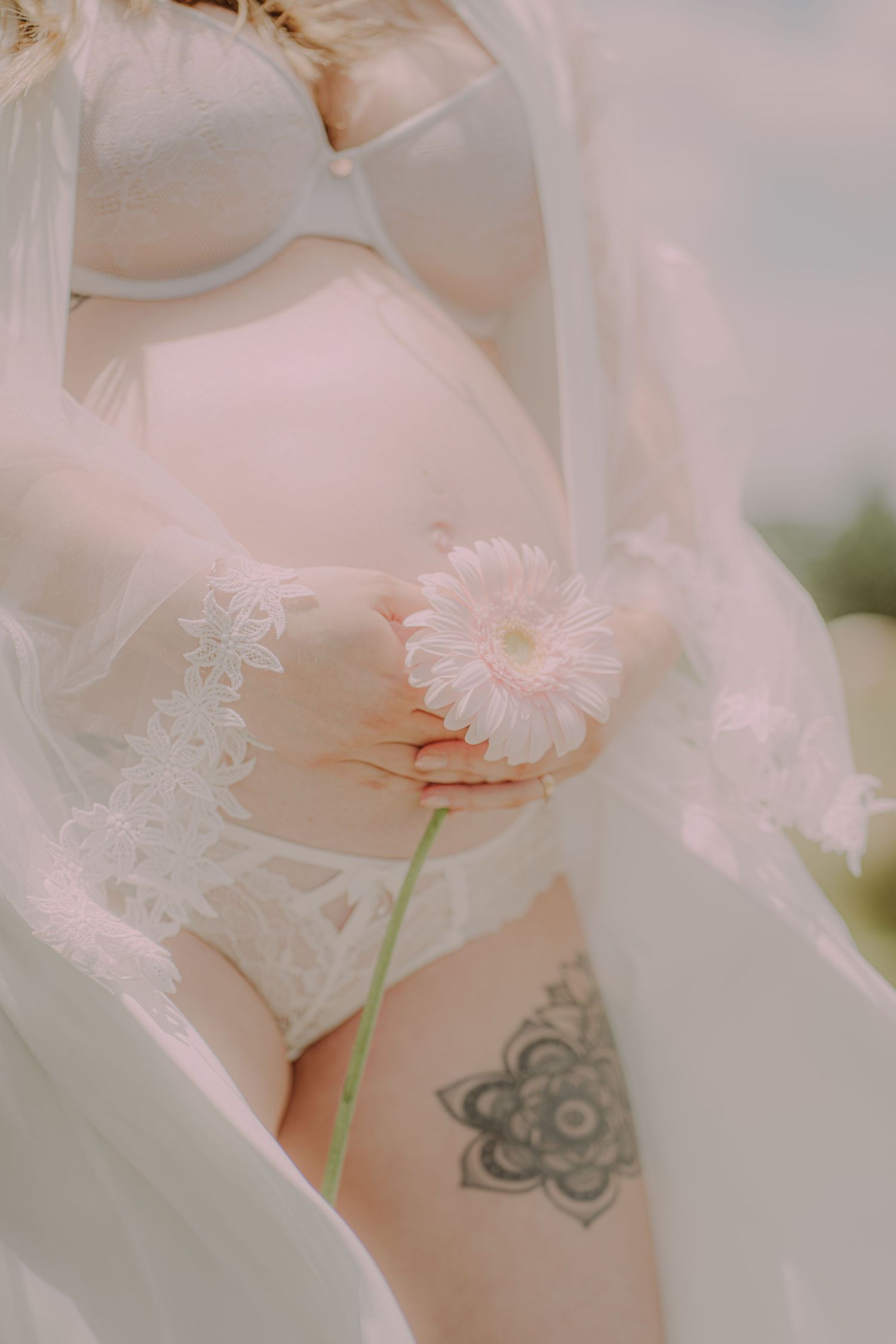Close-up of pregnant woman in white lace lingerie holding pink gerbera daisy with thigh tattoo.