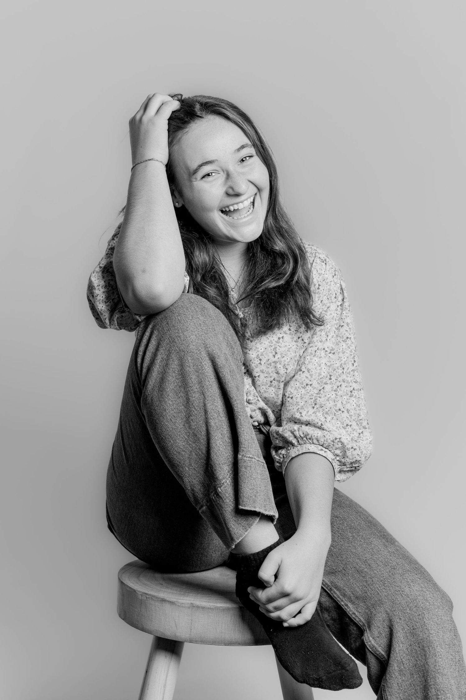 Black and white photo of a laughing teenage girl on a stool, hand in hair, wearing a floral blouse.