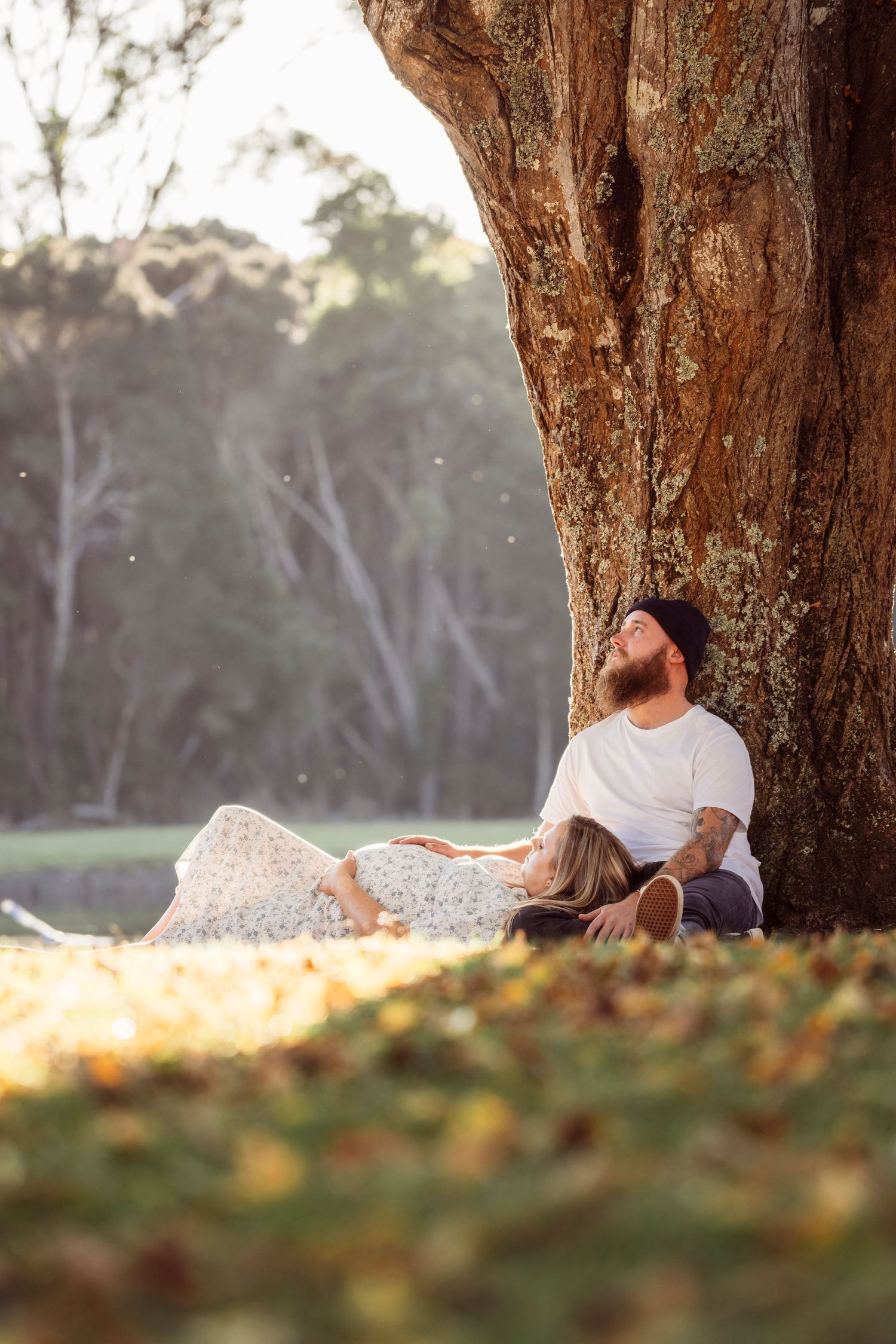 Couple relaxing under a large tree in a sunlit park during a golden hour maternity photoshoot.
