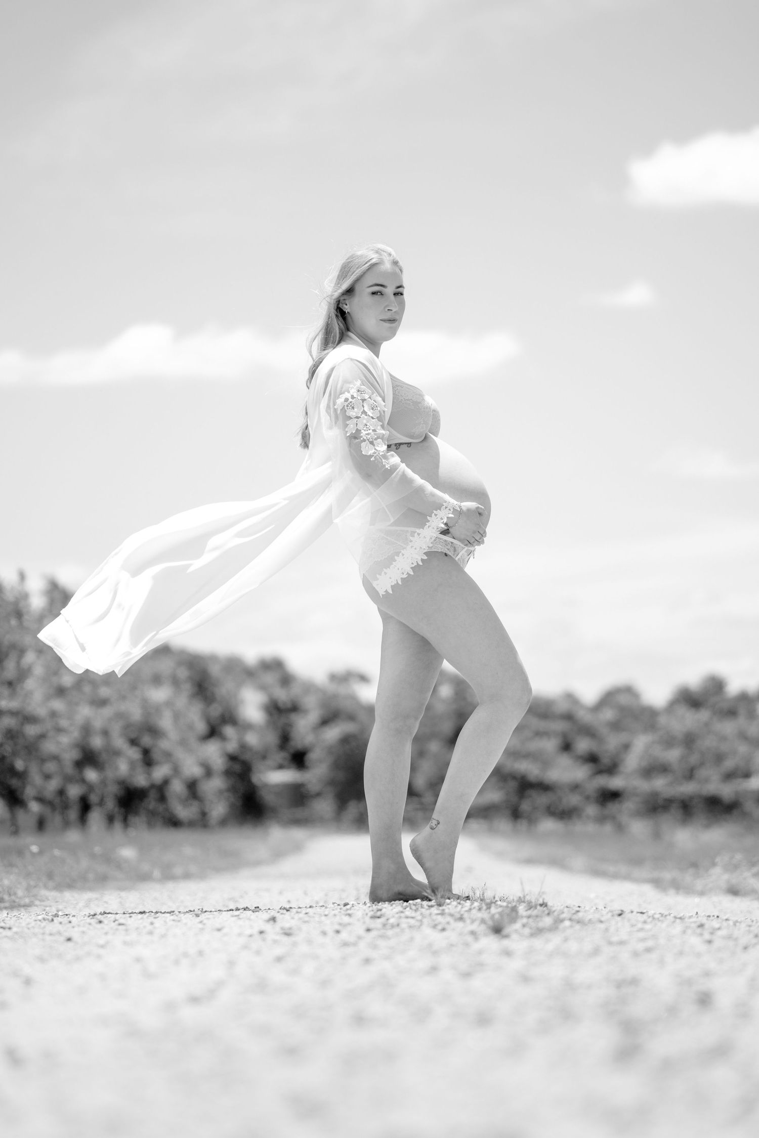 Pregnant woman in white lace robe standing on a gravel path outdoors, black and white maternity photo.