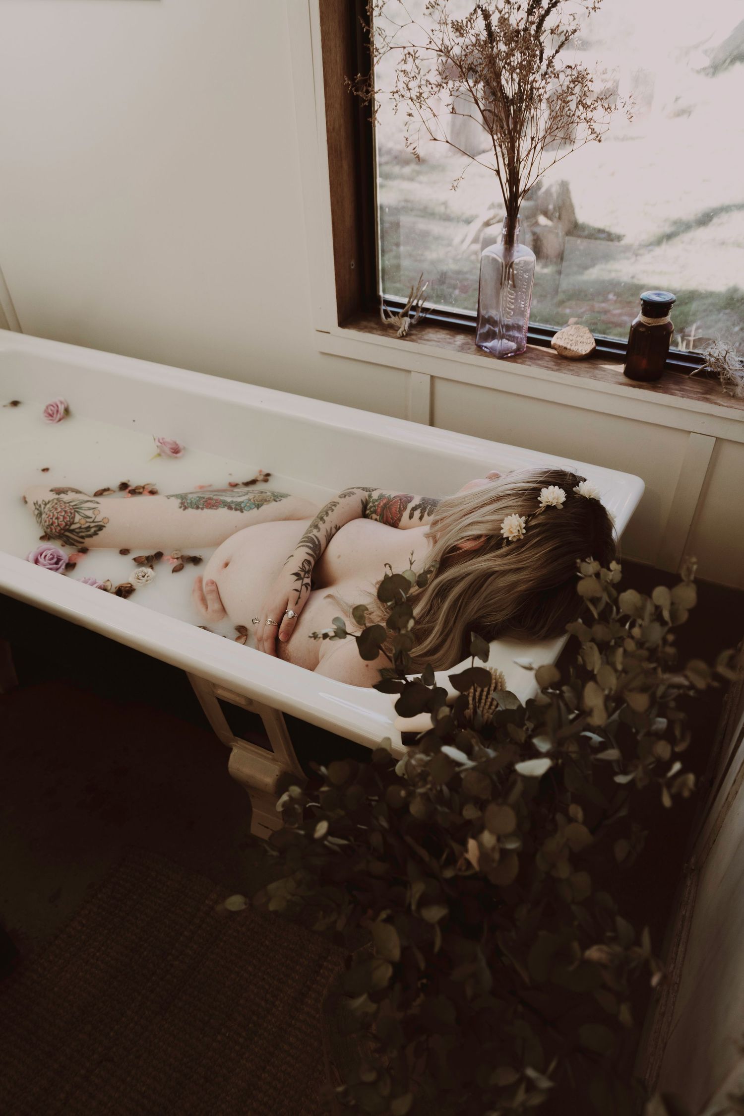 Tattooed pregnant woman relaxing in floral bath with dried botanicals on windowsill nearby.