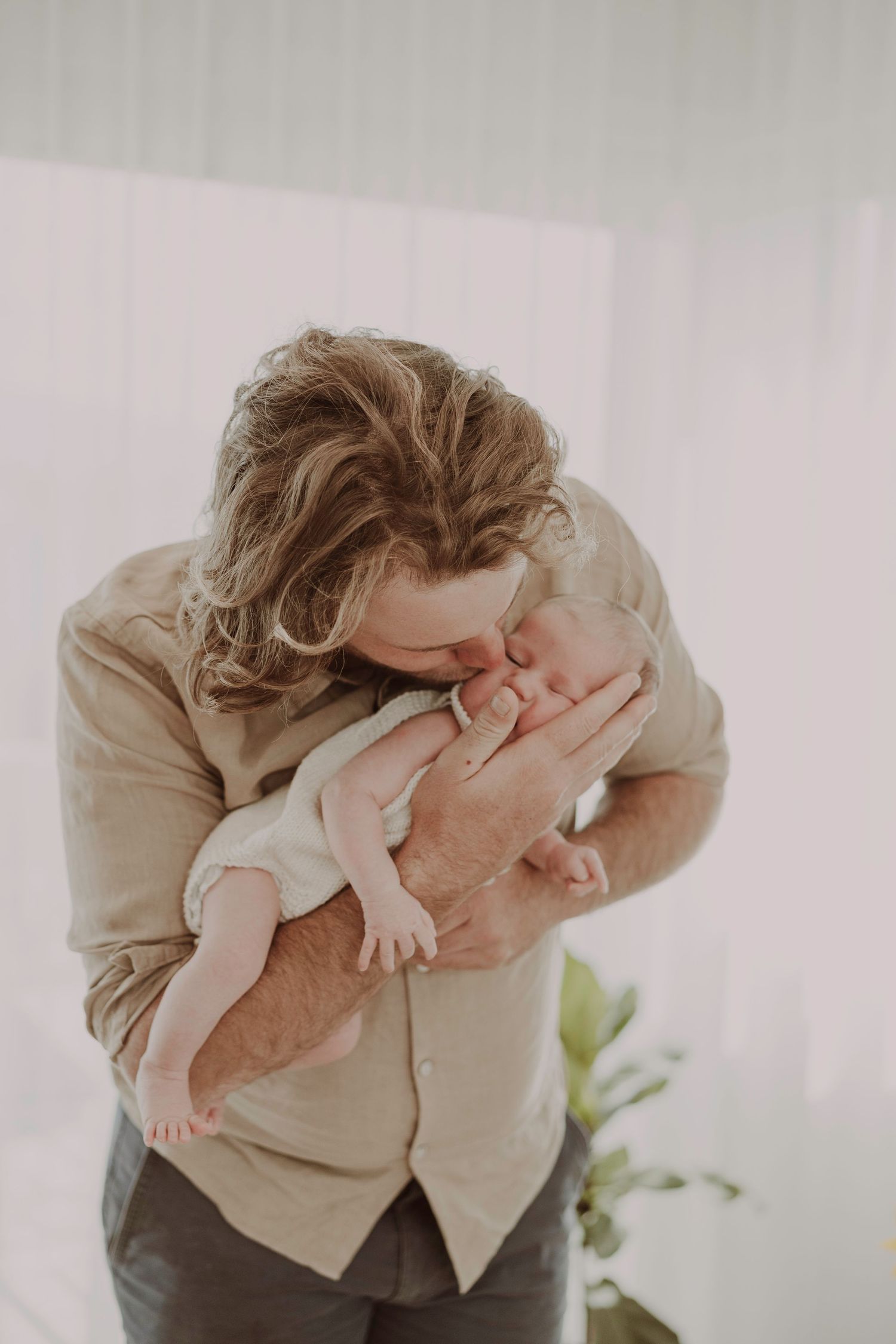 A father with wavy hair gently kisses his newborn baby held in his arms in a bright room.