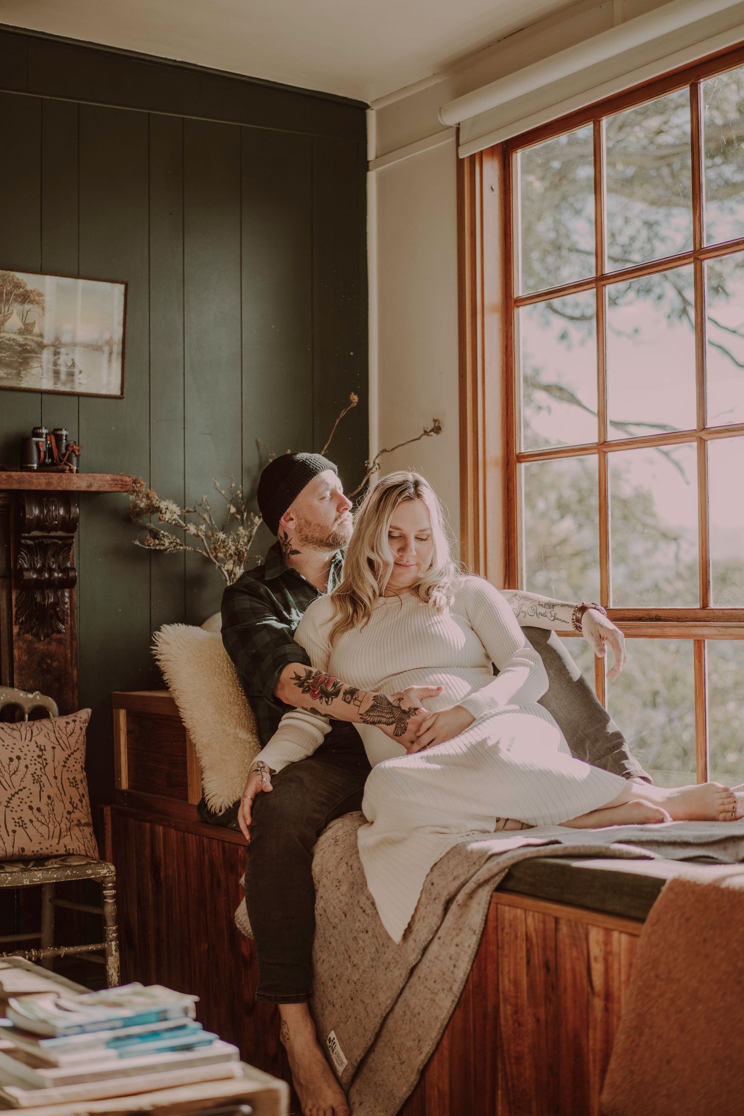 Tattooed groom and bride sitting together on vintage daybed by large window in cozy room.