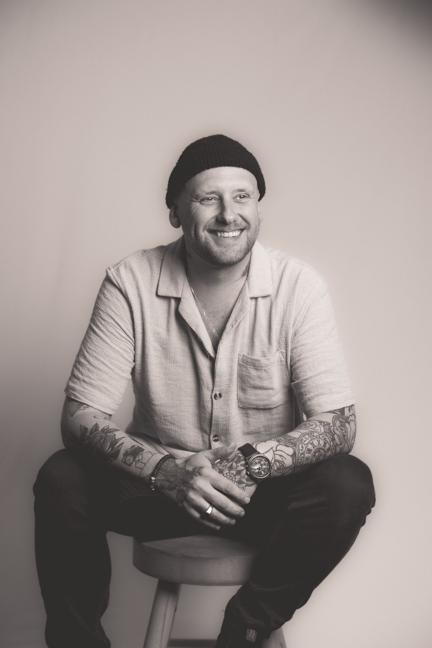 Black and white studio portrait of smiling tattooed man in beanie sitting on stool.