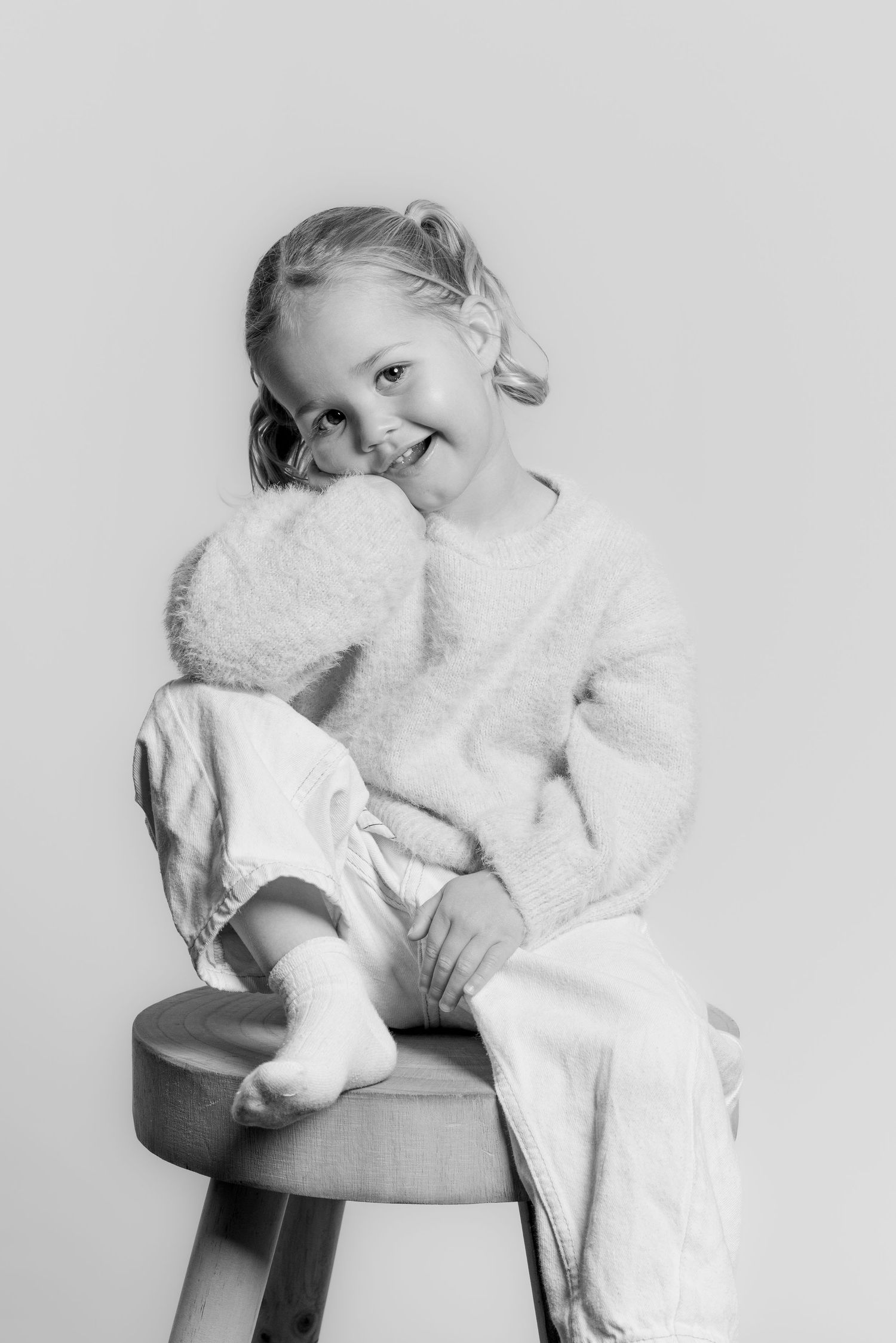 Black and white portrait of a smiling young girl sitting on a stool, hugging a stuffed animal toy.
