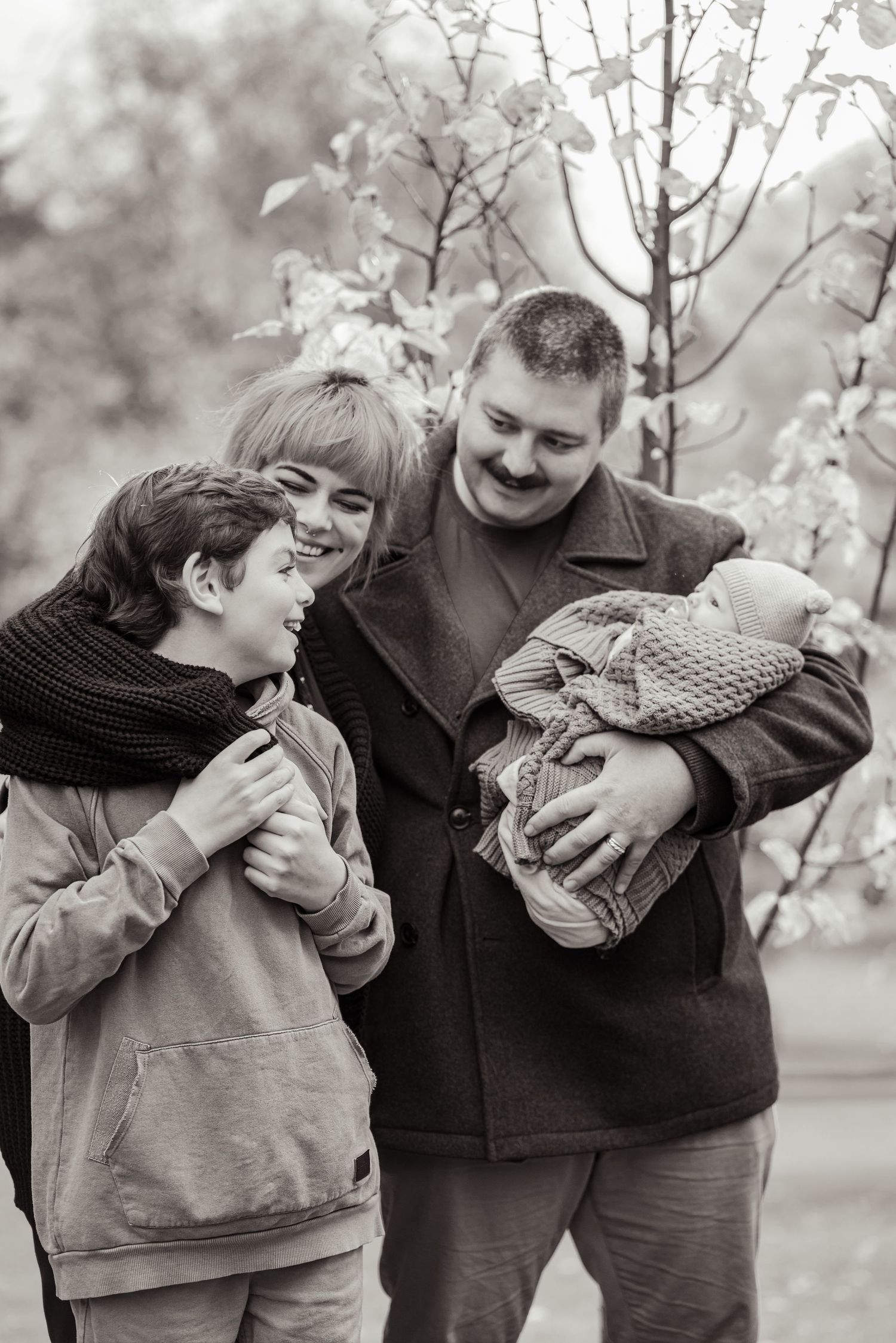 Black and white family portrait with parents and child admiring newborn baby outdoors.