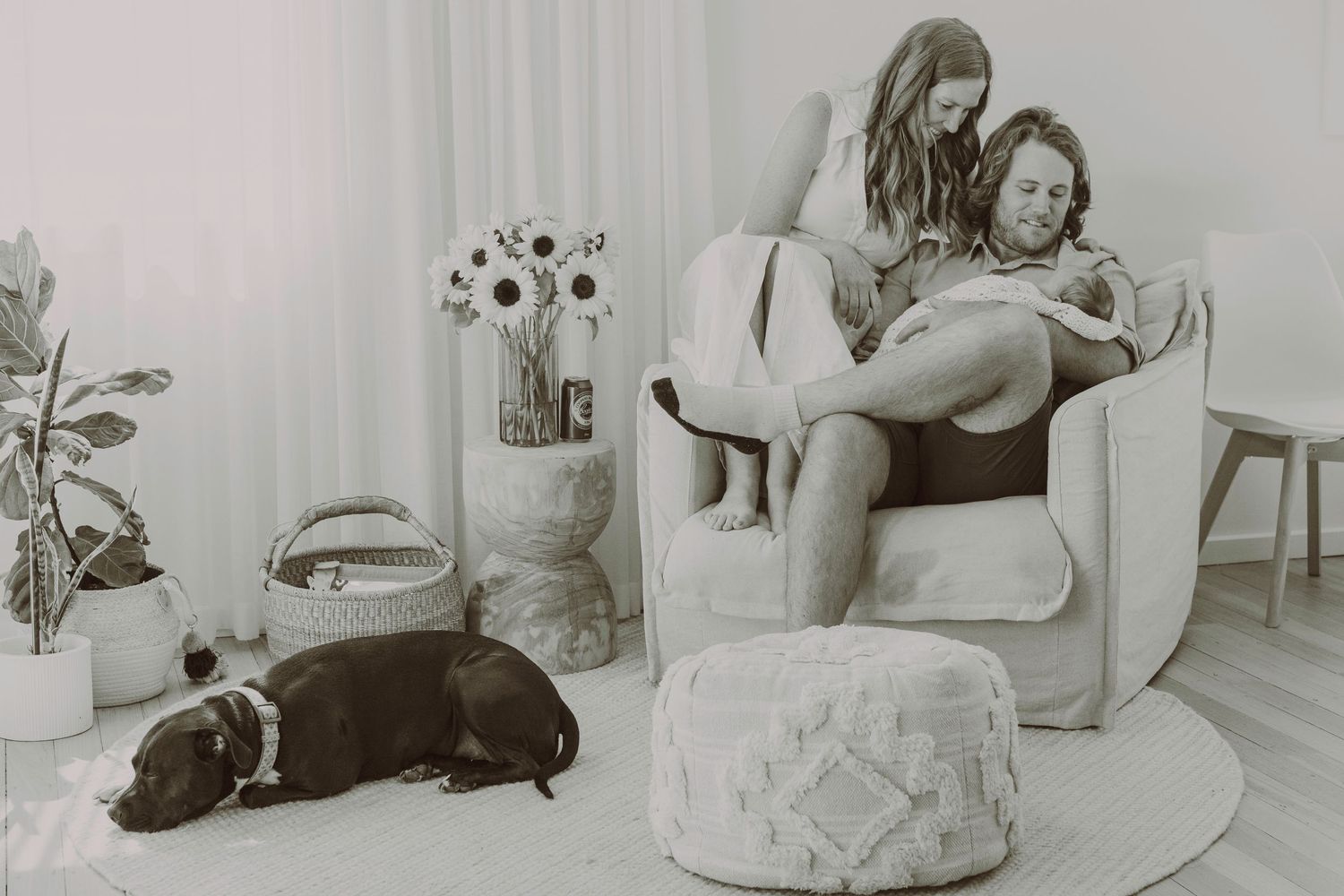 New parents sit together in a cozy nursery chair looking at their newborn while their dog rests nearby.