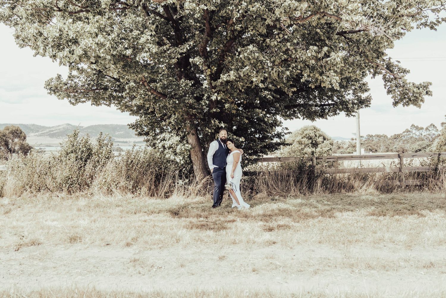 A bride and groom stand beneath a large tree in a scenic outdoor wedding portrait.