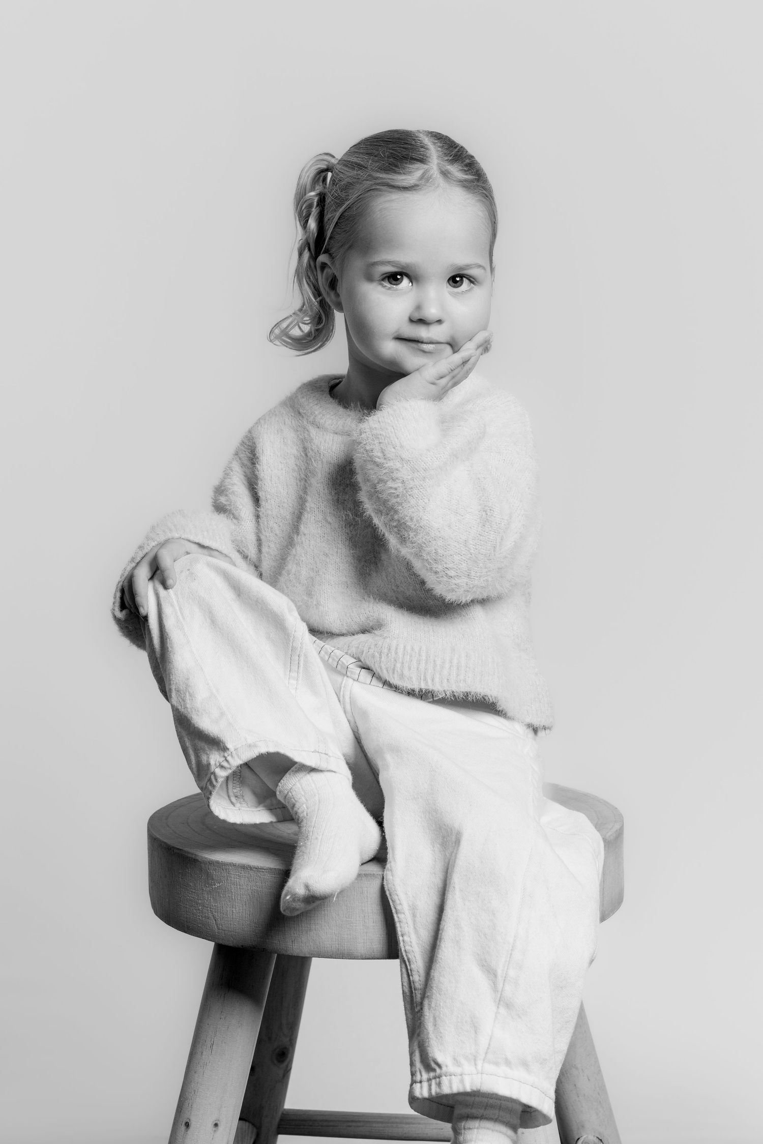 Black and white photo of a little girl sitting on a wooden stool, resting her cheek on her hand.