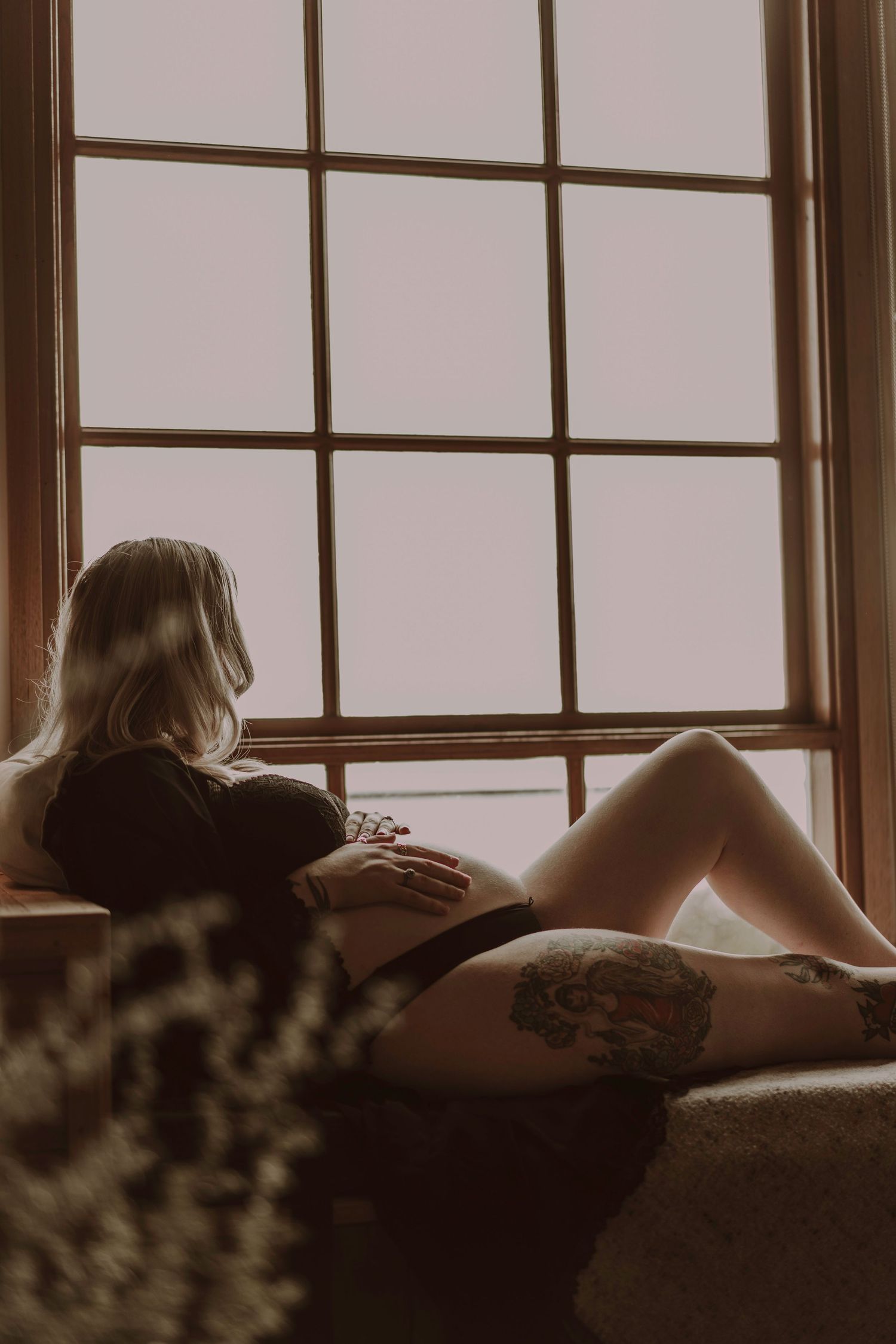 Pregnant tattooed woman reclining on sofa gazing out large window in warm natural light.