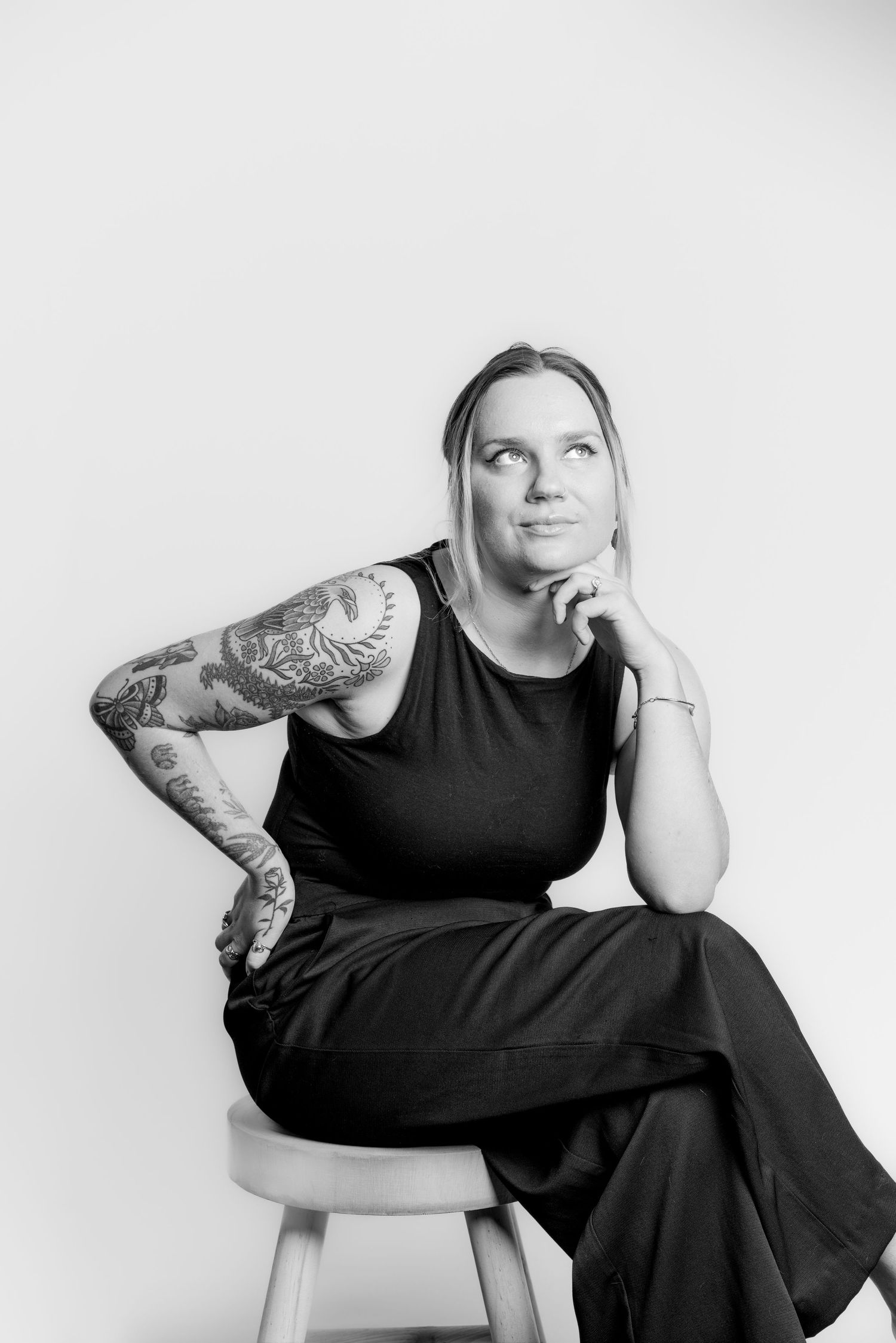 Black and white portrait of thoughtful tattooed woman in black outfit seated on stool.