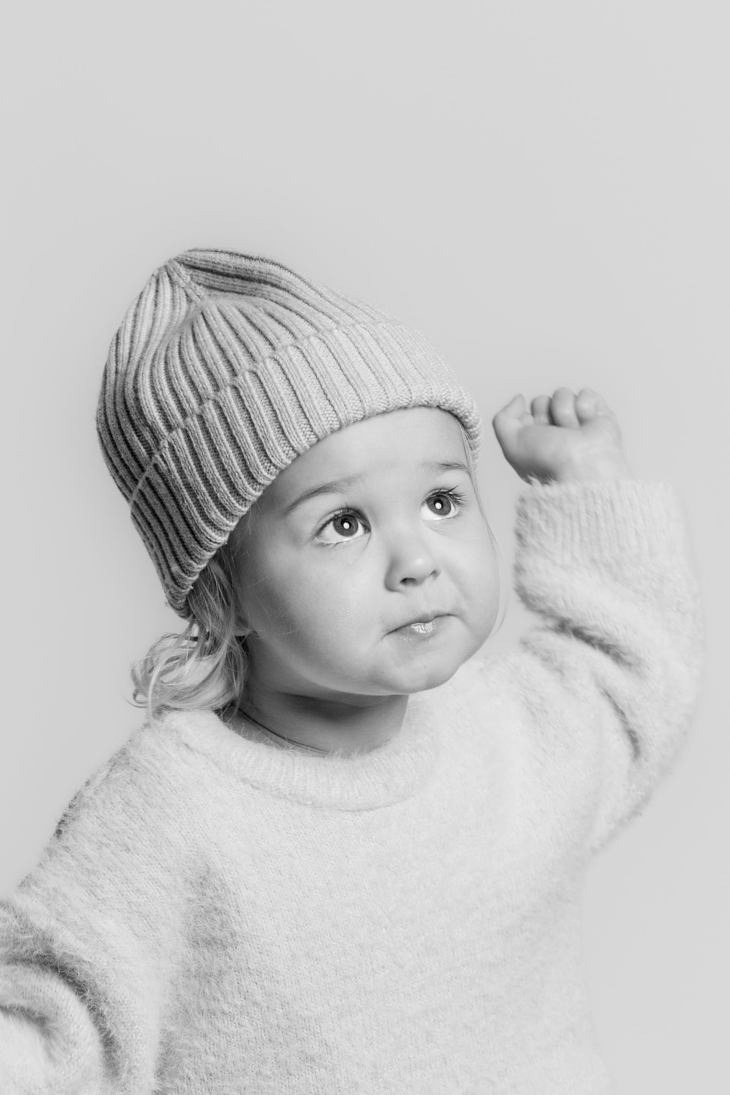 Black and white portrait of a young toddler in a knit beanie and fluffy sweater raising one fist with a curious expression.