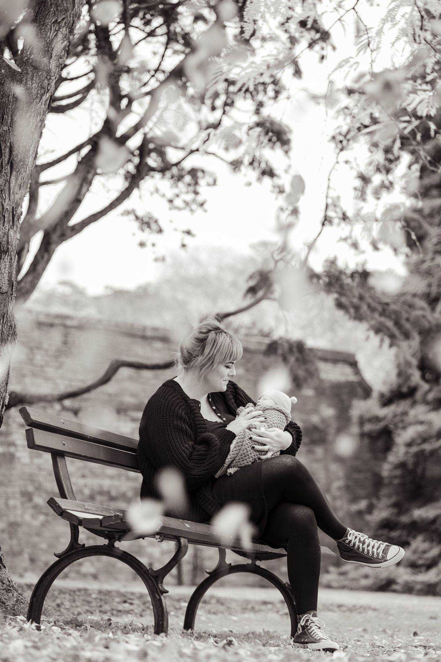 Black and white photo of mother sitting on park bench holding newborn baby under tree.
