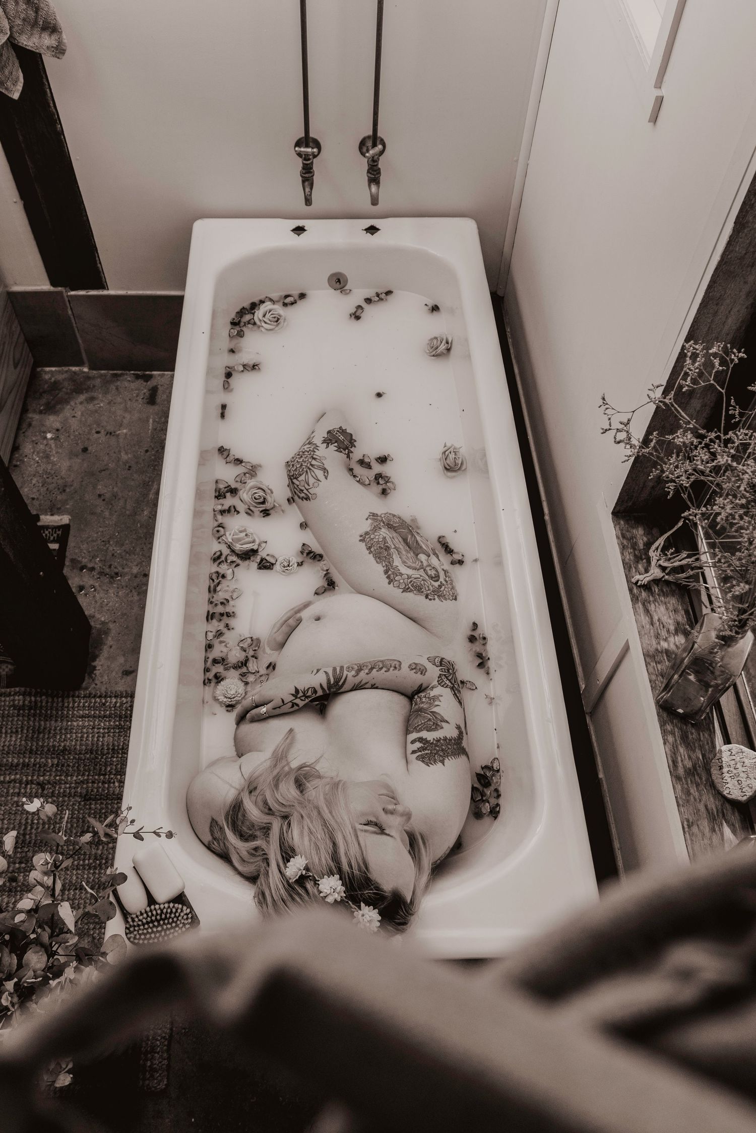 Overhead black and white photo of a tattooed pregnant woman in a floral milk bath.
