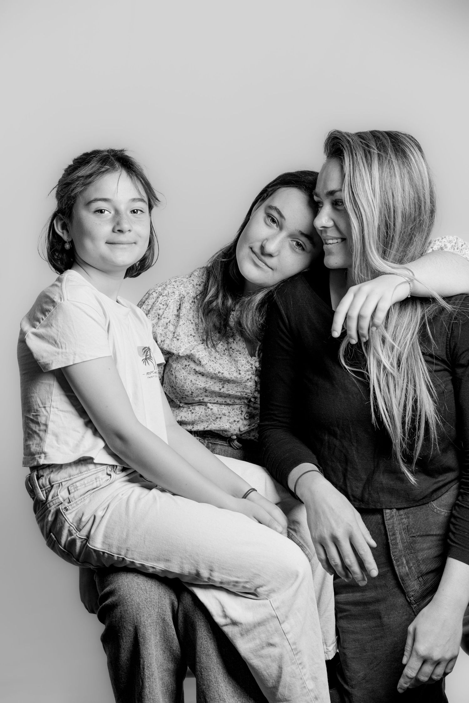 Three females lean together smiling in a candid black and white studio portrait.