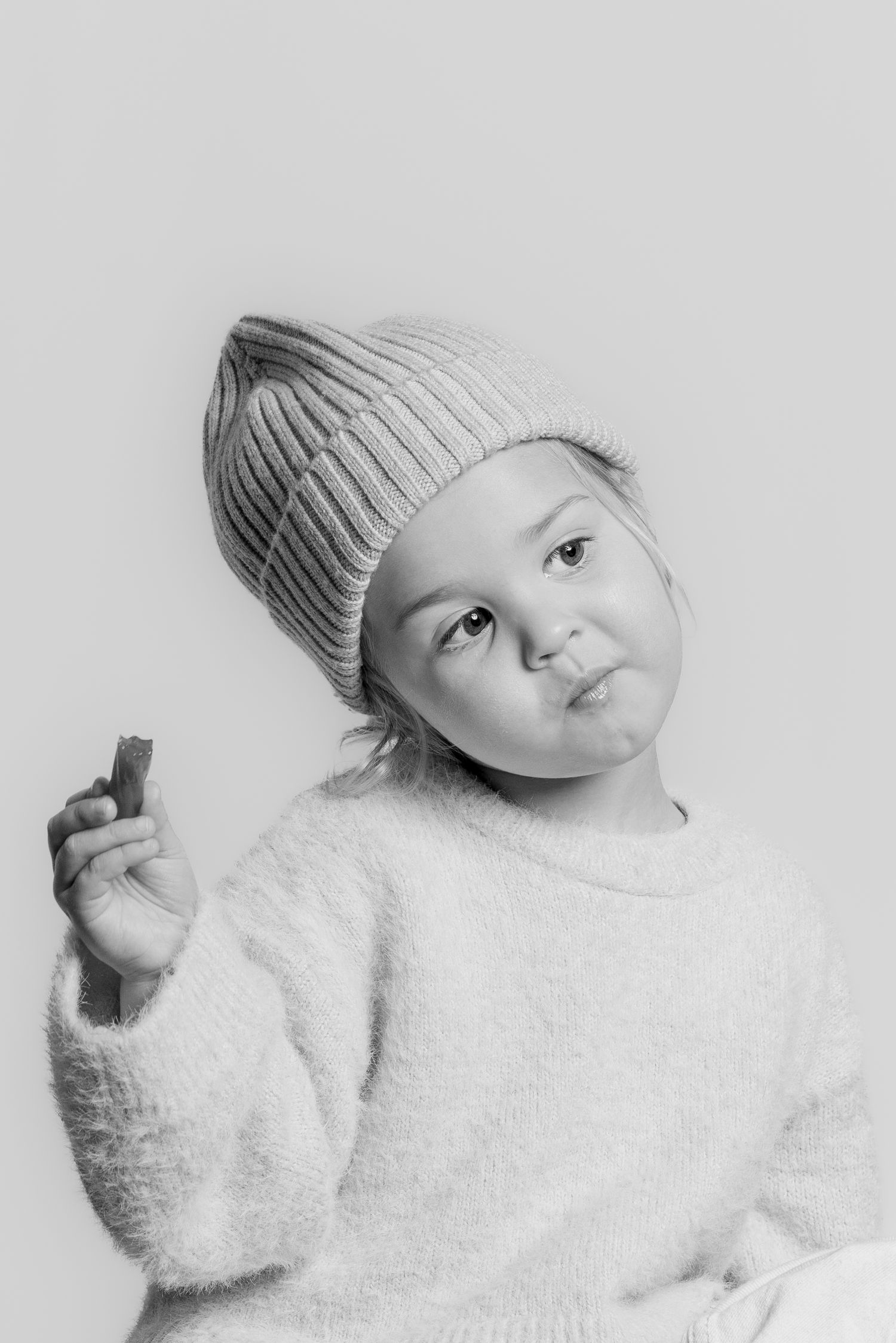 Black and white portrait of a young child wearing a ribbed knit beanie and fluffy sweater holding a small object.