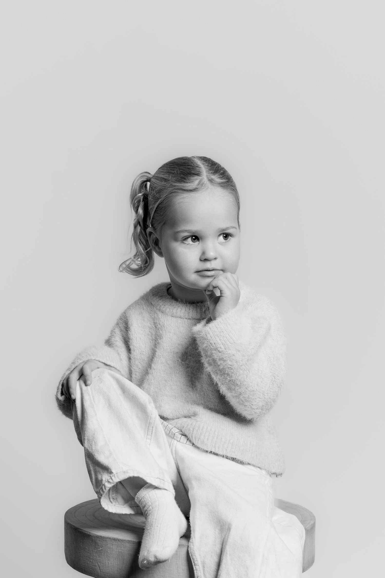 Black and white portrait of a thoughtful young girl sitting on a stool in a cozy knit sweater and wide-leg pants.