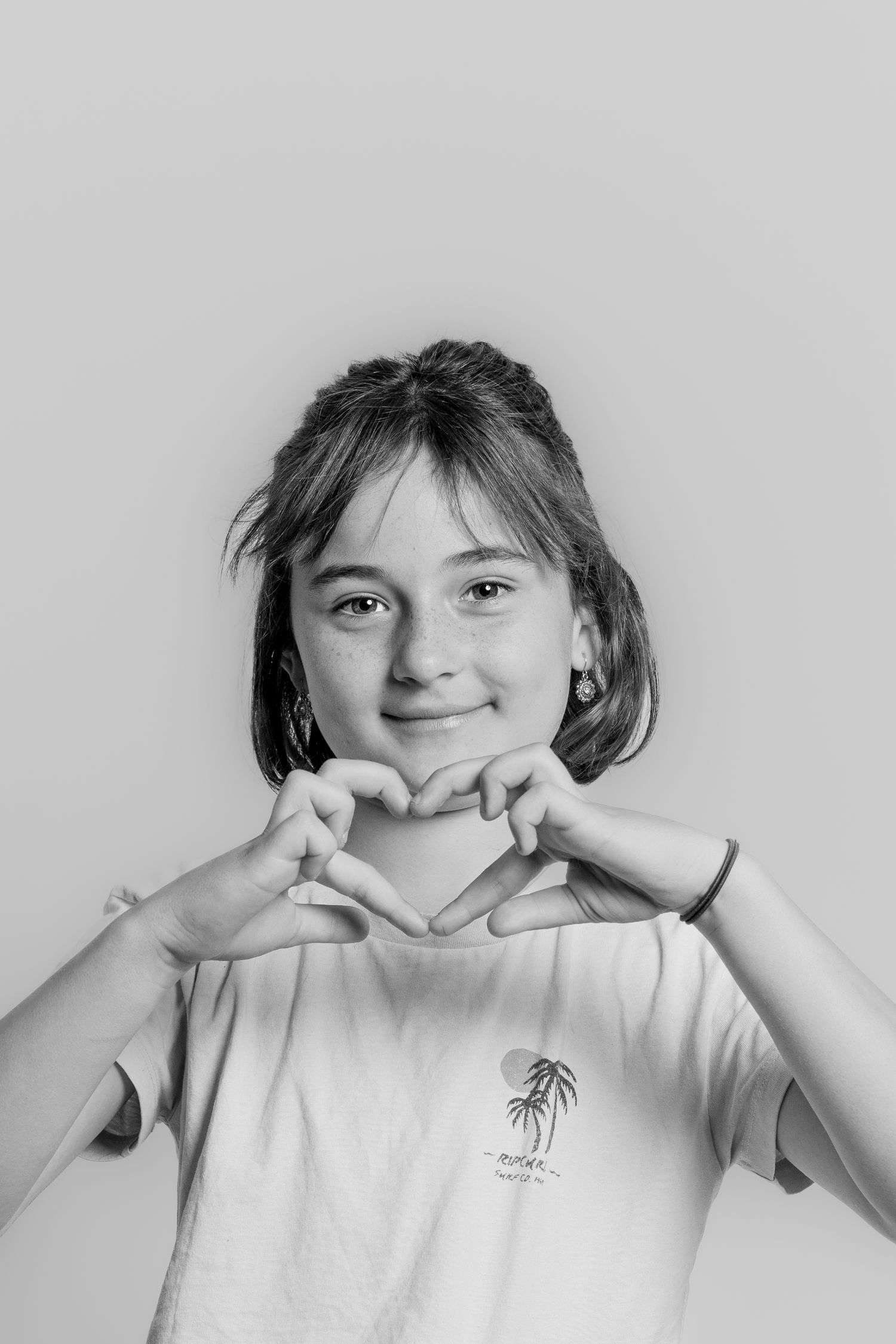 Black and white photo of a young girl making a heart shape with her hands, wearing a white t-shirt.