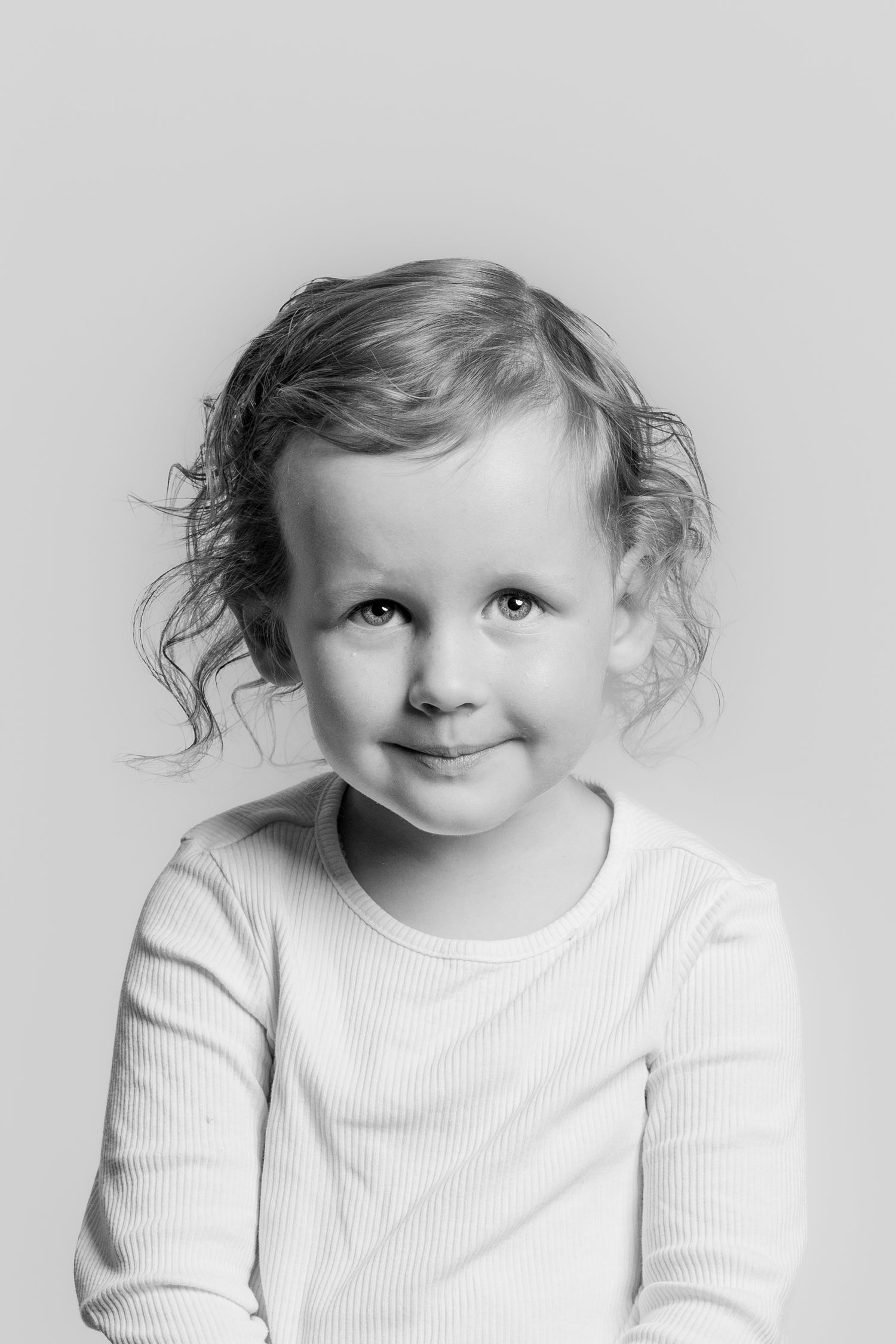 Black and white close-up portrait of a smiling toddler girl with curly hair in a white top.