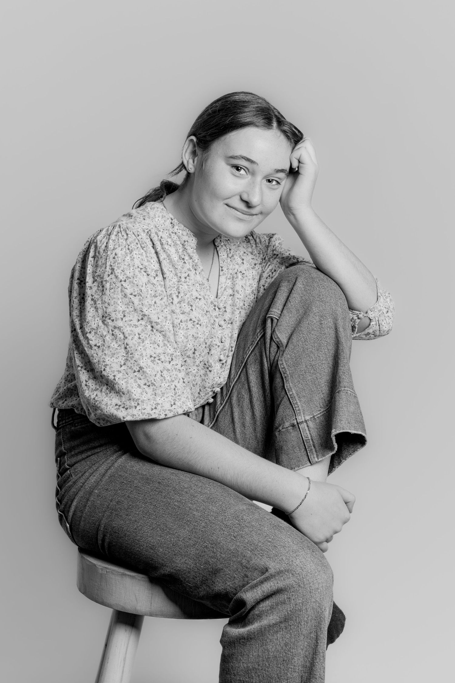 Black and white portrait of a teenage girl sitting on a stool, resting head on hand, smiling softly.