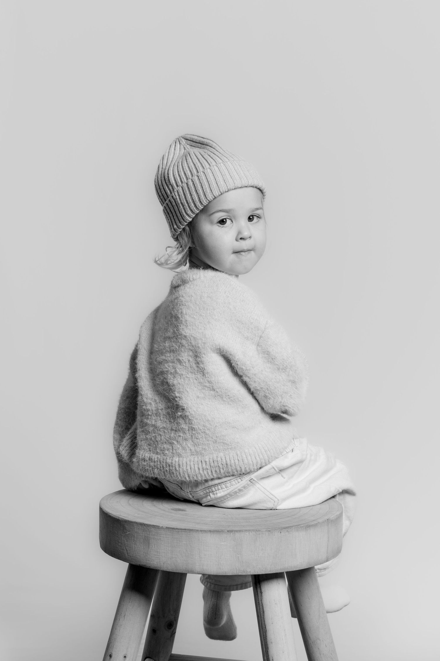 Black and white photo of a smiling toddler in a knit beanie sitting on a wooden stool.