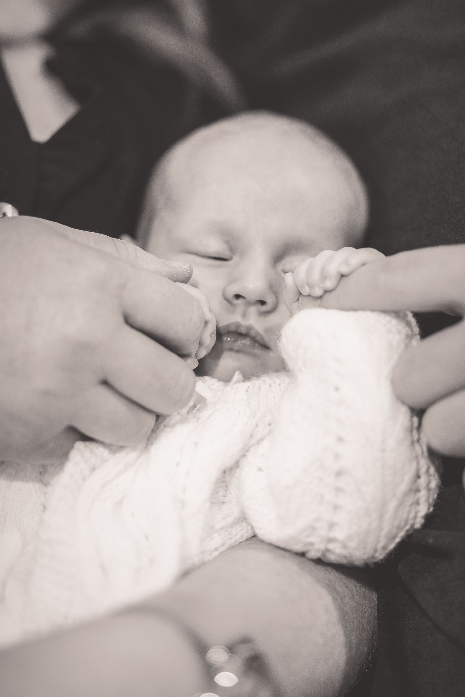 Black and white close-up of sleeping newborn baby in knit outfit holding parent finger.