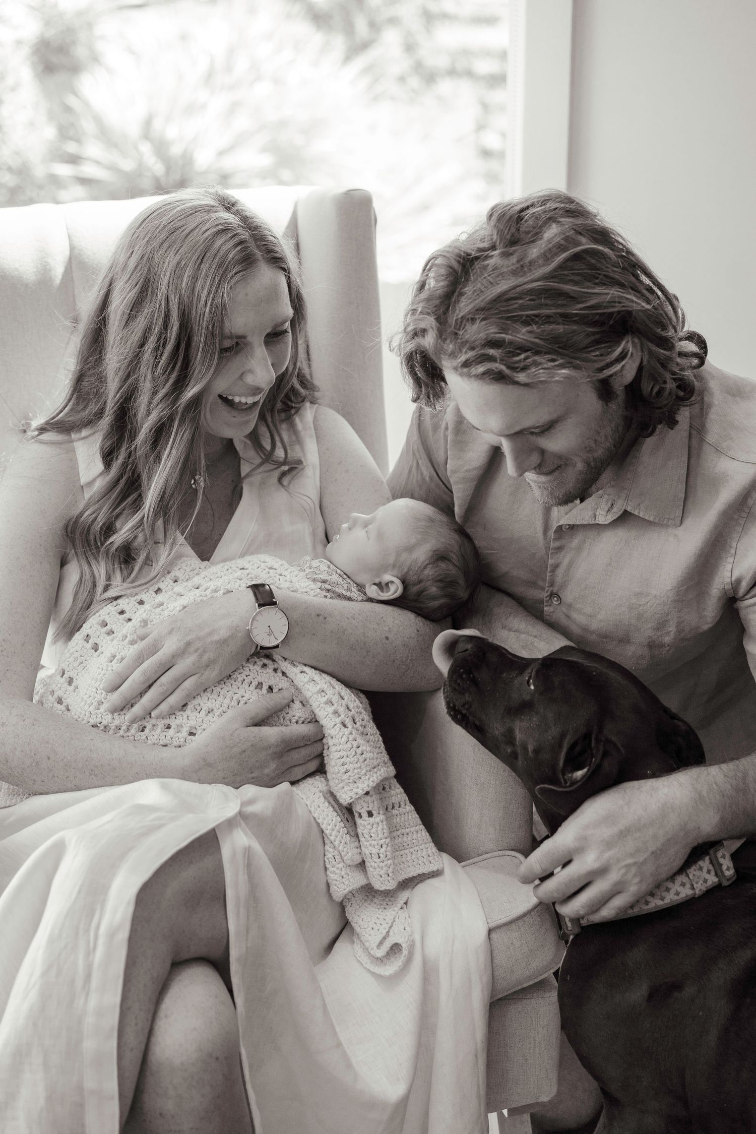 Happy new parents smile at their sleeping newborn baby while their black dog looks on in black and white photo.