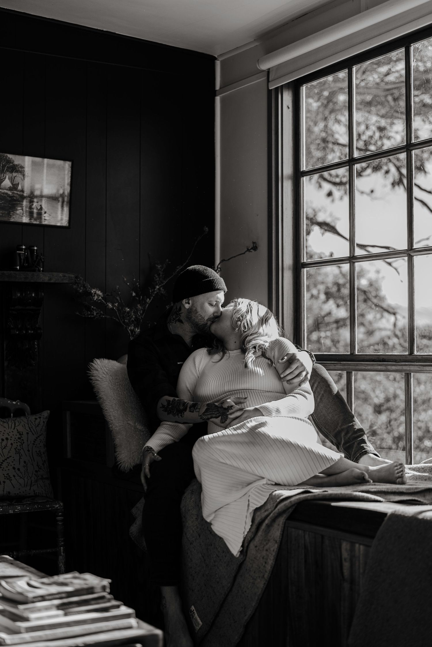 Black and white photo of couple sharing a tender kiss by a large window indoors.
