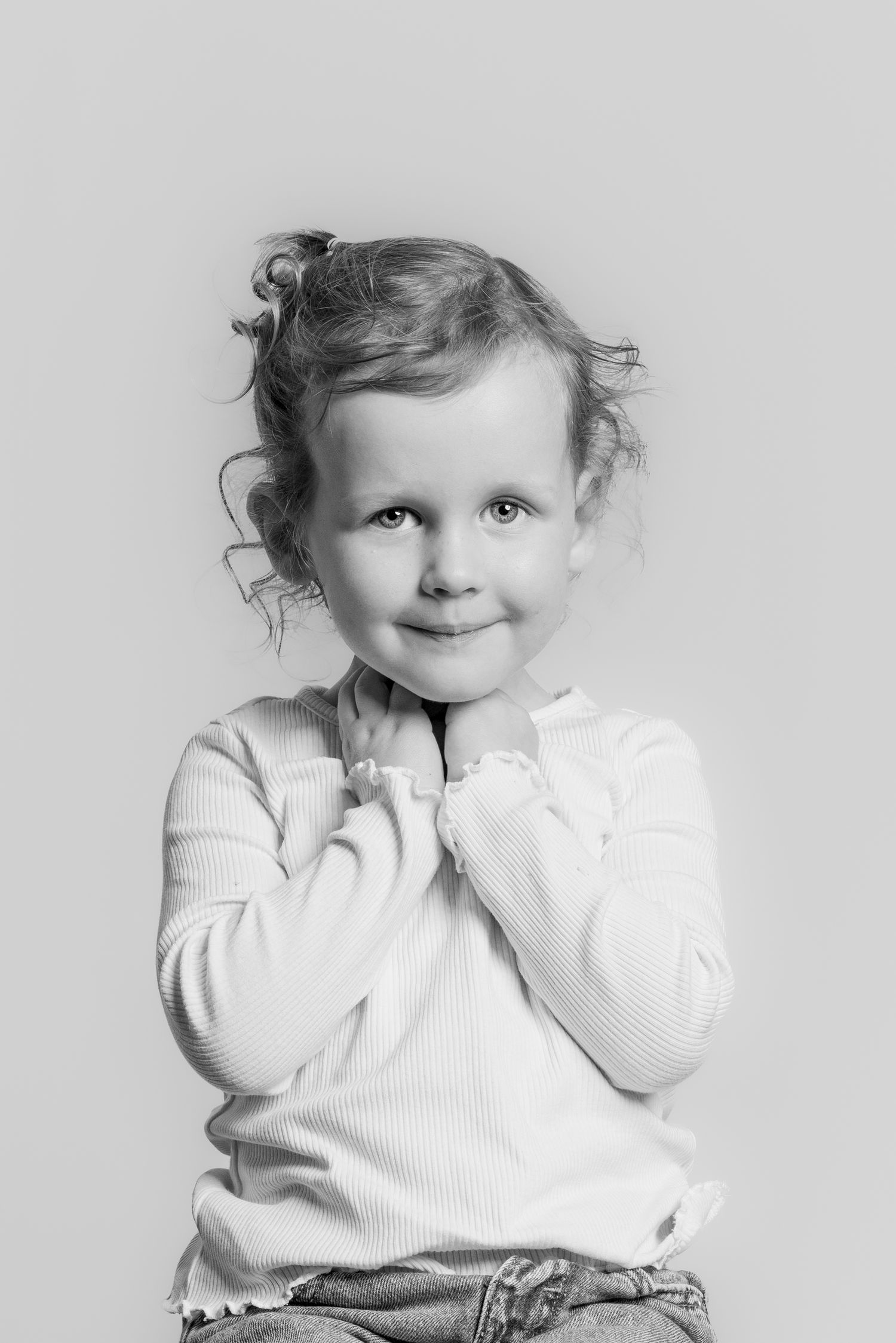 Black and white portrait of a sweet toddler girl with curly hair, hands clasped under her chin.
