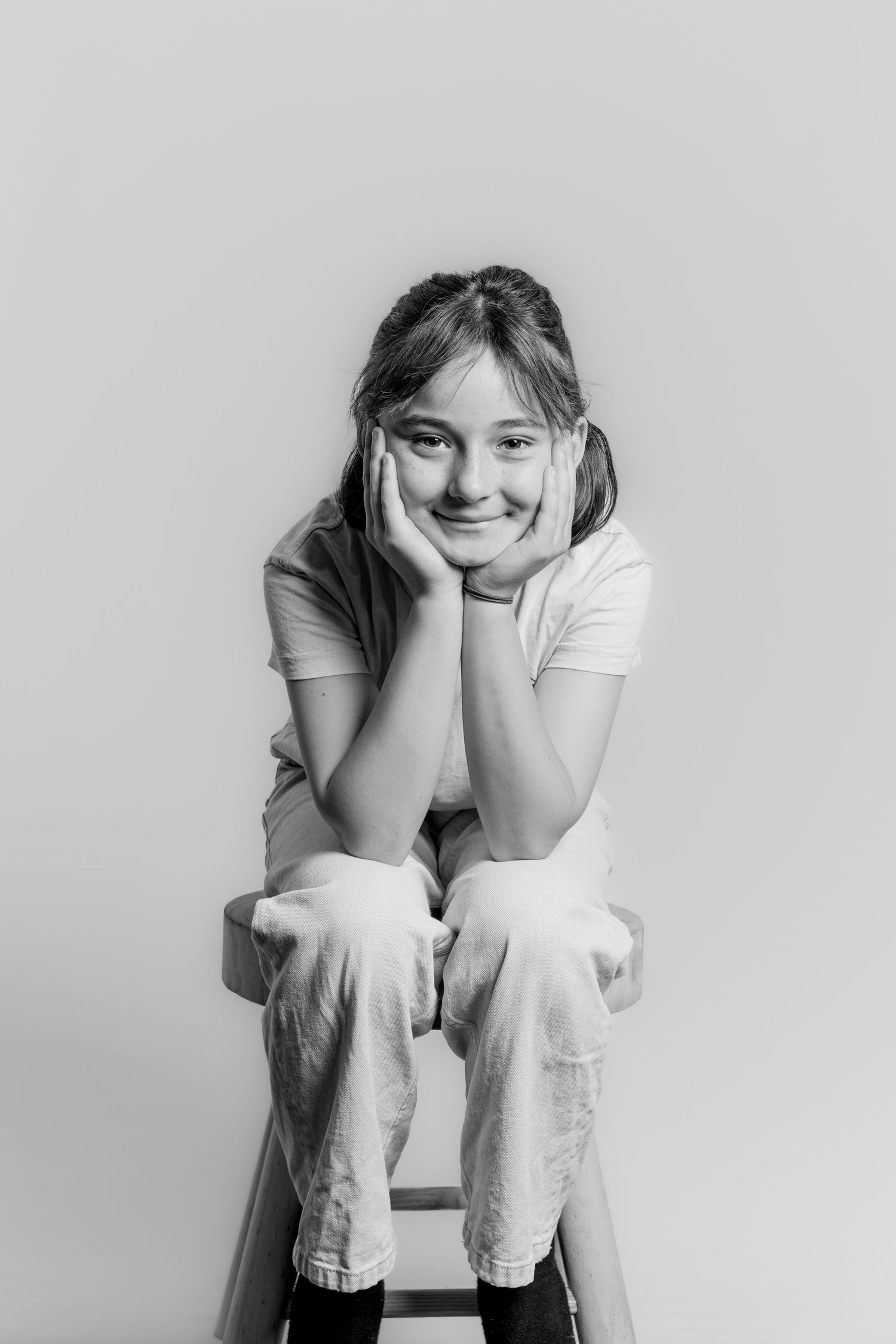 Black and white photo of a young girl sitting on a stool with hands on cheeks, smiling at camera.