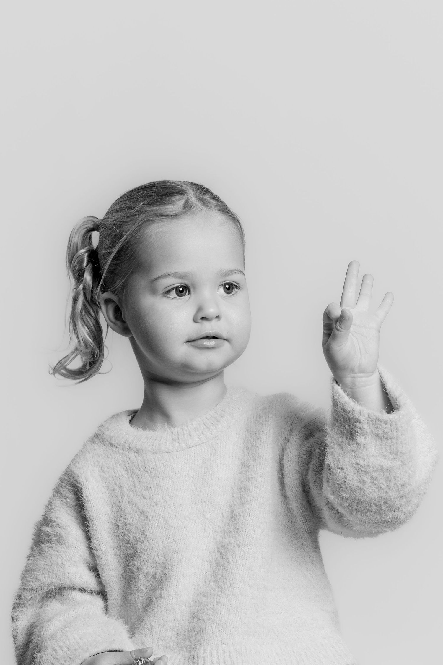Black and white portrait of a young girl holding up three fingers, wearing a fluffy sweater.