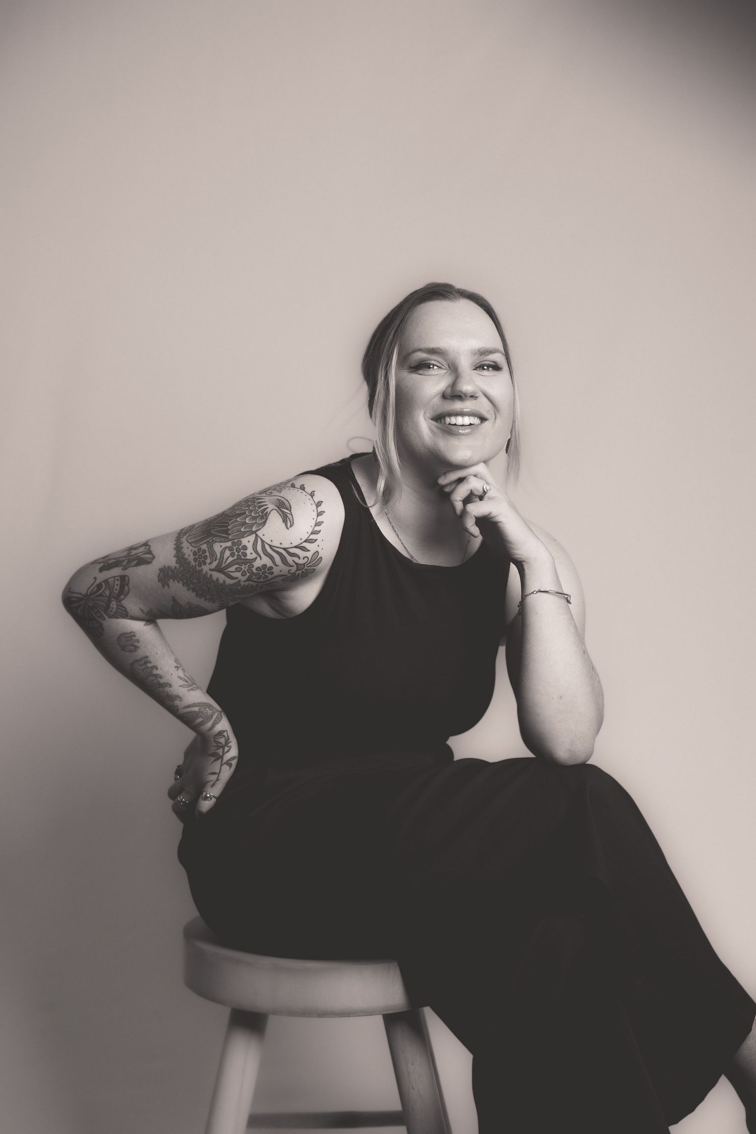Black and white studio portrait of smiling tattooed woman in black outfit on stool.