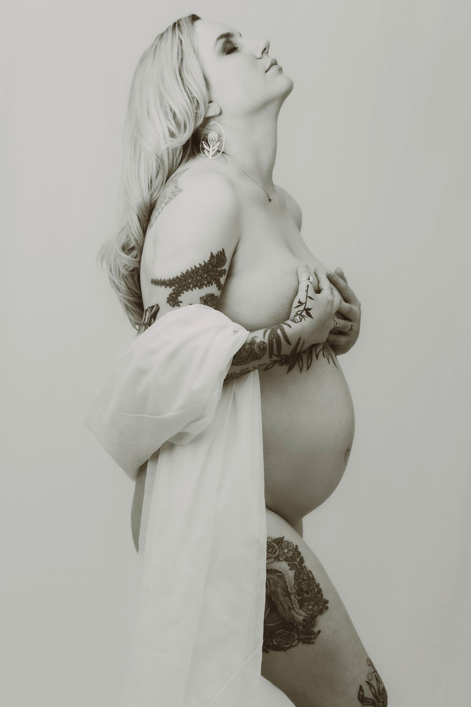Artistic sepia maternity photo of a tattooed pregnant woman in a flowing white robe.