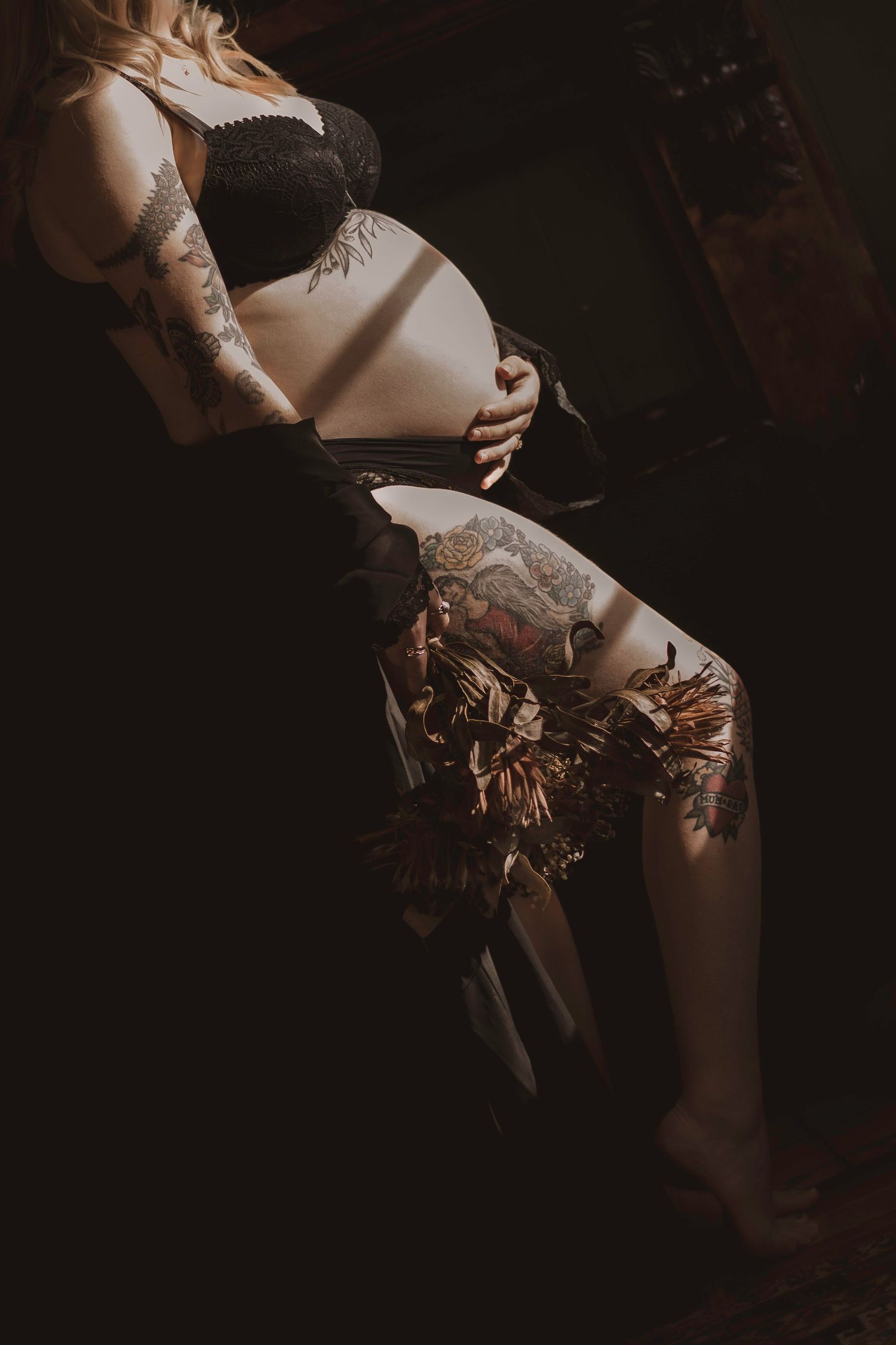 Pregnant woman with tattoos in black lingerie cradling baby bump in moody dark setting.