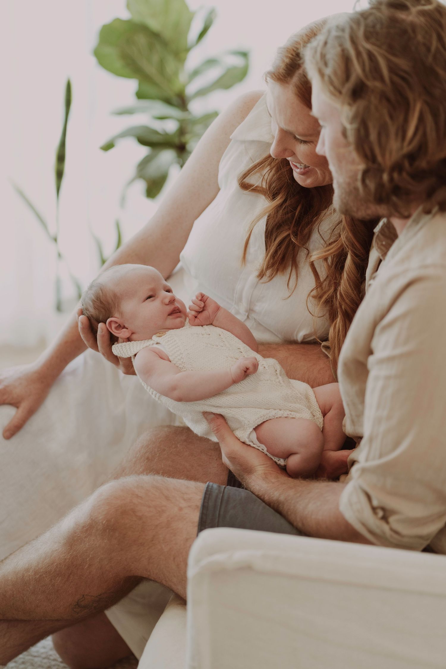 New parents sitting on a white couch lovingly looking at their alert newborn baby in a white onesie.