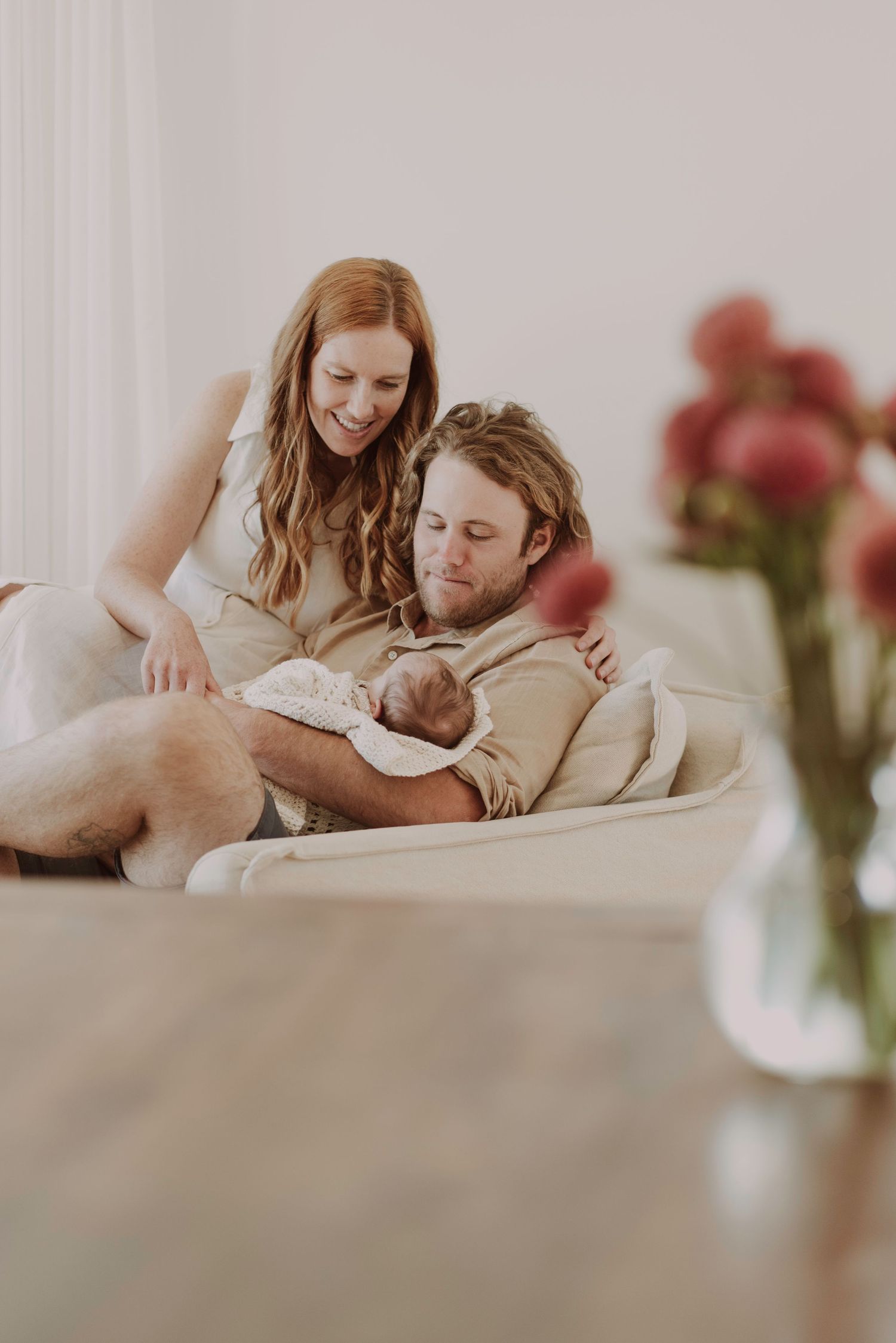 A couple smiles lovingly at their newborn baby while seated on a couch.