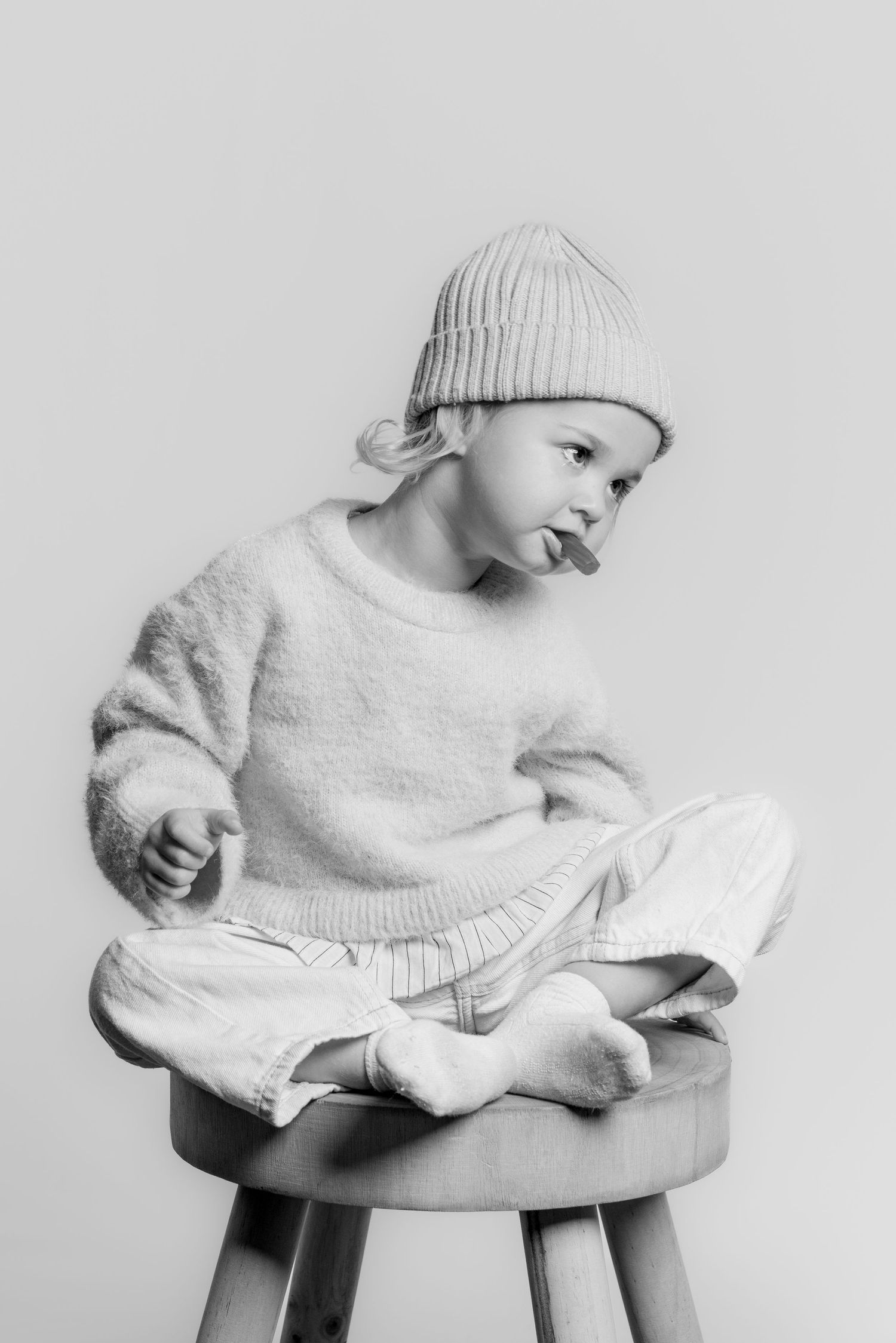 Black and white portrait of a young child sitting cross-legged on a wooden stool, wearing a knit hat.