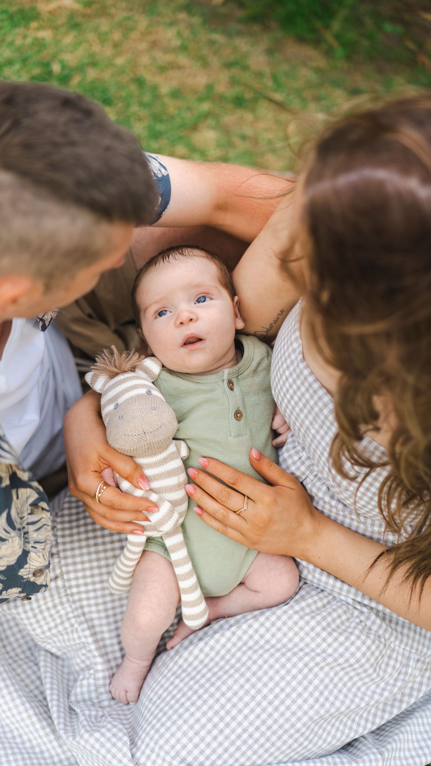 Parents lovingly holding their wide-eyed baby with a striped toy in a garden.