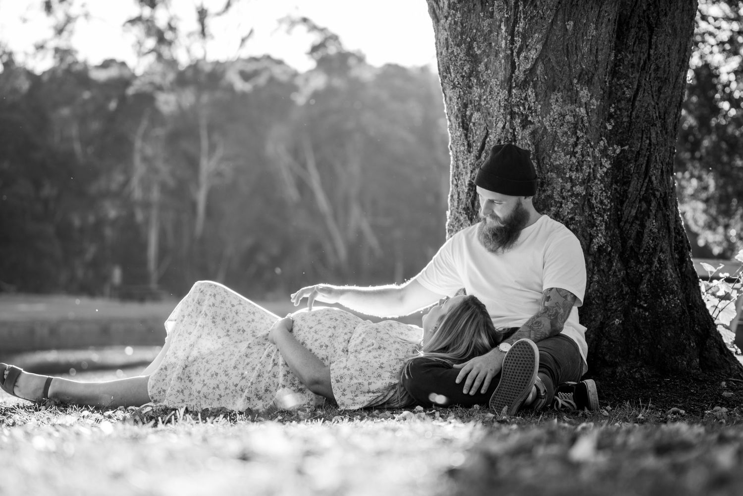 Bearded man in beanie sitting under tree with pregnant partner in floral dress, black and white.