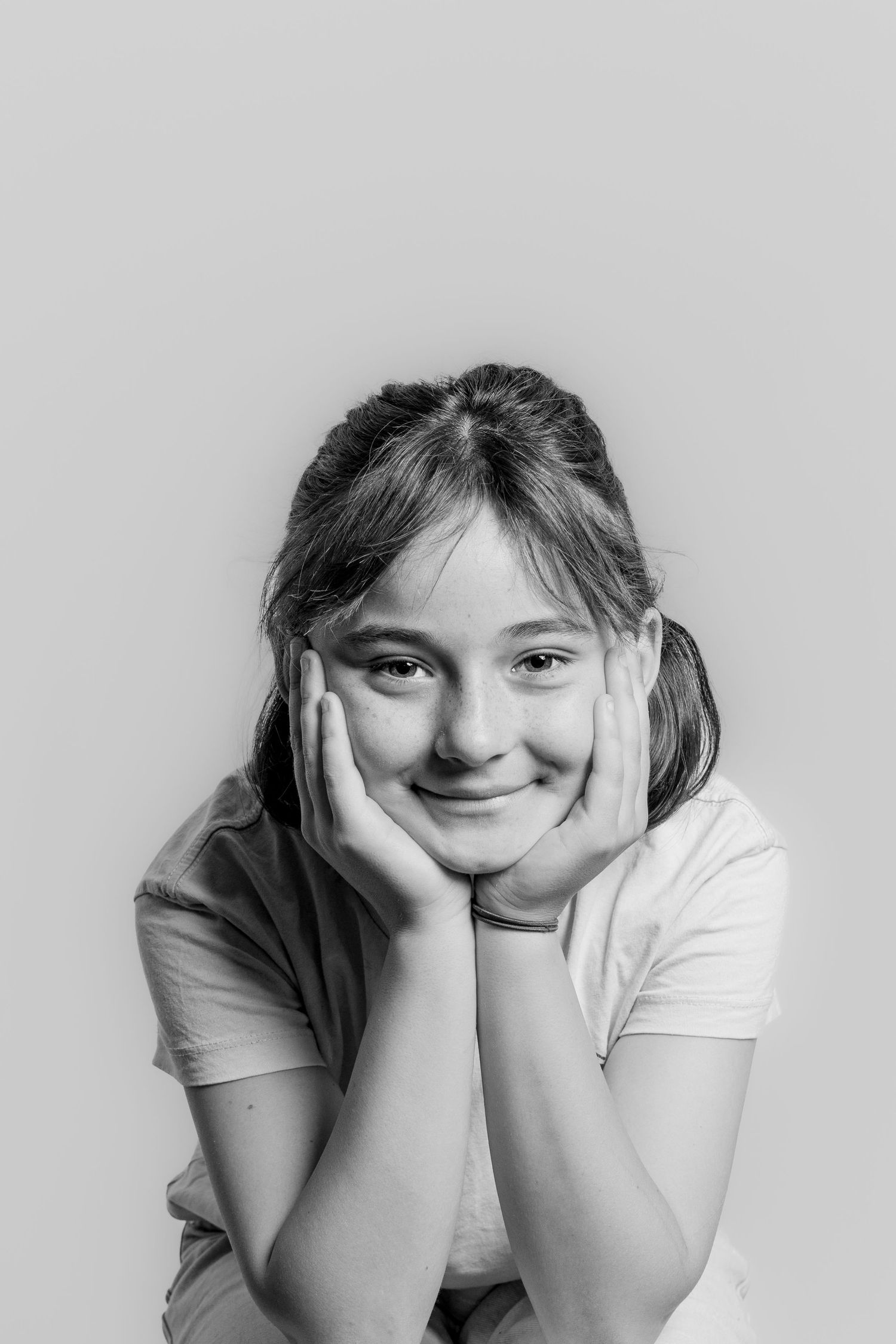 A young girl rests her chin on her hands, smiling softly in a black and white portrait.