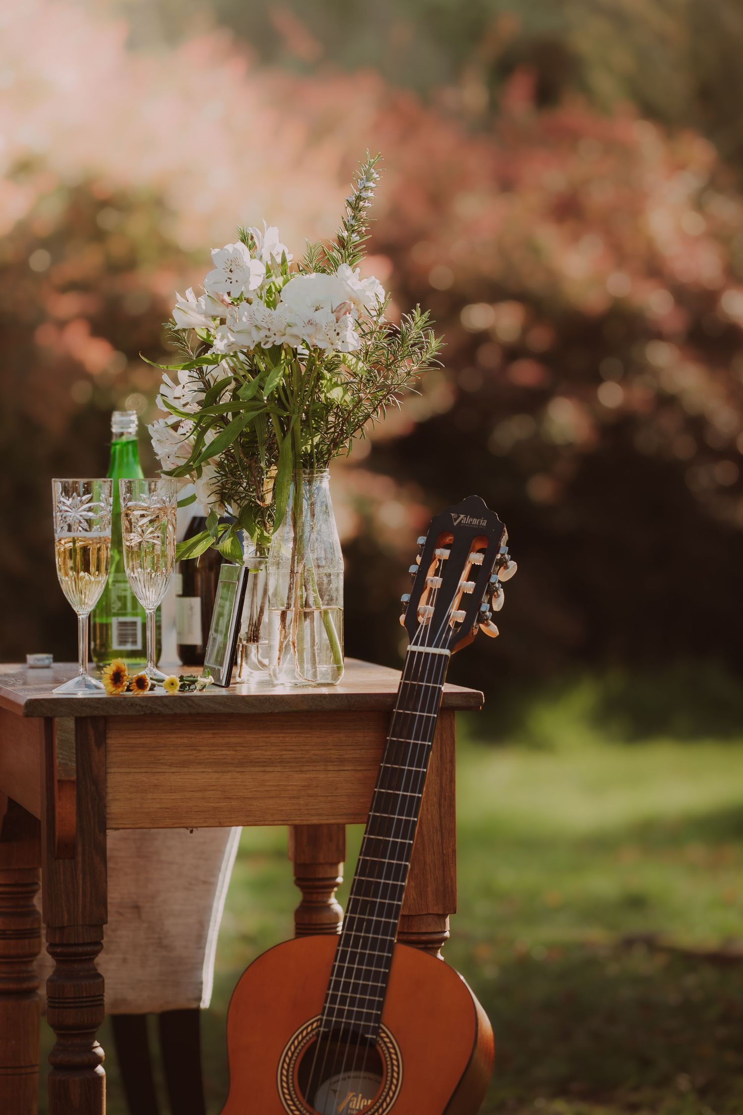 Acoustic guitar leaning against a wooden table with white floral arrangement, champagne glasses, and wine bottles in a garden setting.