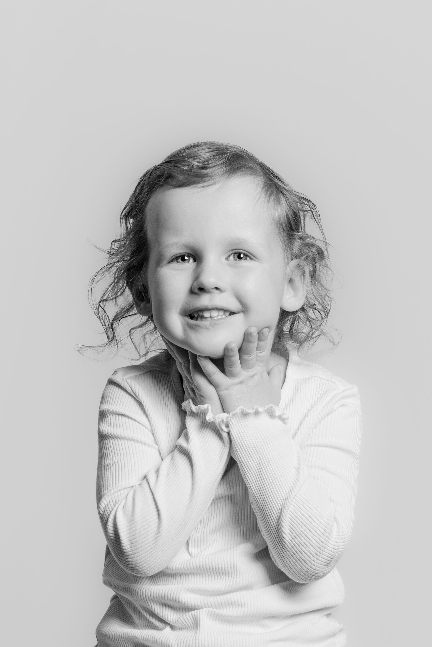 Black and white portrait of a cheerful curly-haired girl in a white top, hands near her face.