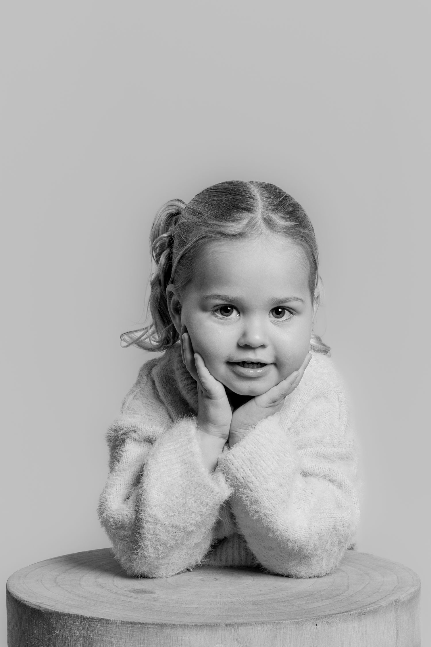 Black and white portrait of a young girl leaning forward on a wooden stool, resting her face in her hands.