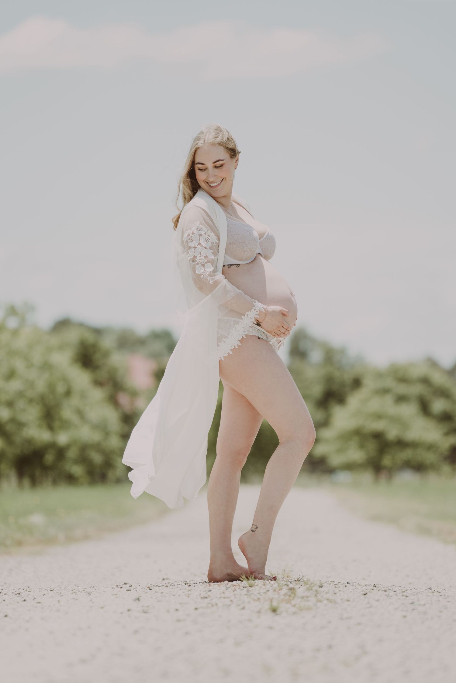 Smiling pregnant woman in white lace robe cradling her baby bump on a sunny gravel path.
